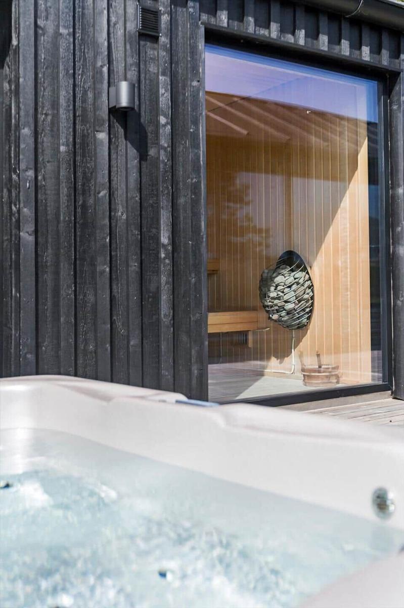 Eine Sauna mit Holzwand und runden Steinobjekt hinter Glasfenster. Im Vordergrund Whirlpool mit Wasser.
