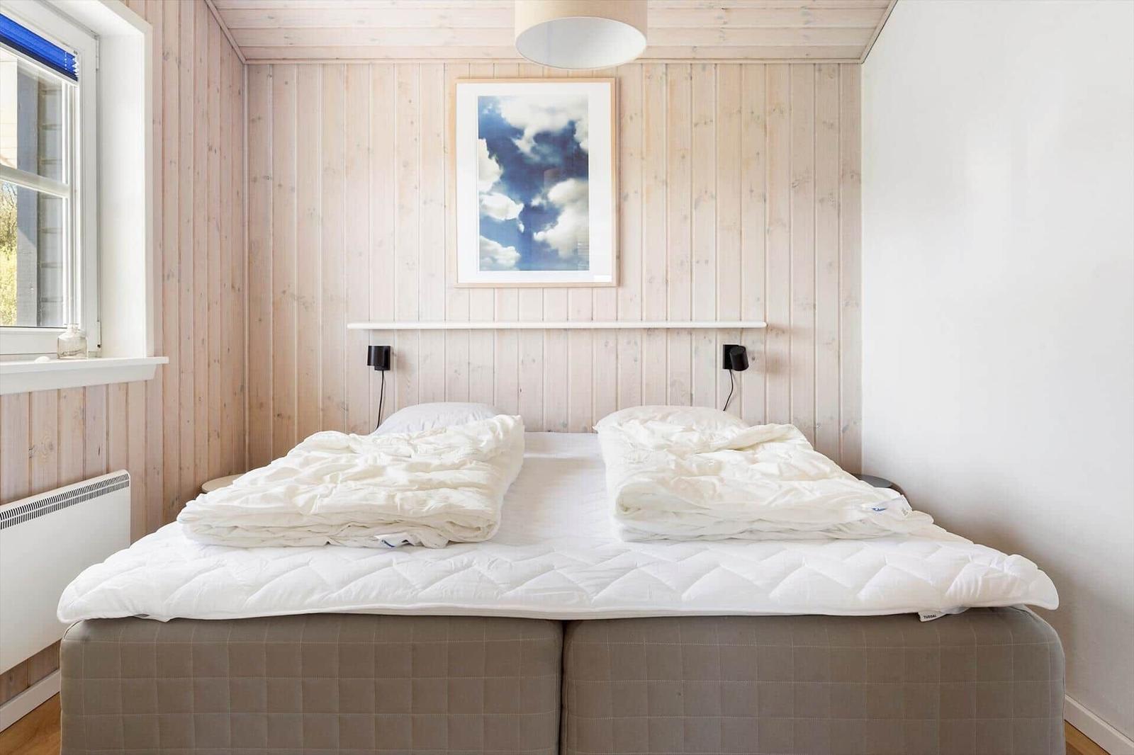 Schlafzimmer mit Doppelbett, Holzwand und Fenster. Decke und Kissen sind weiß.