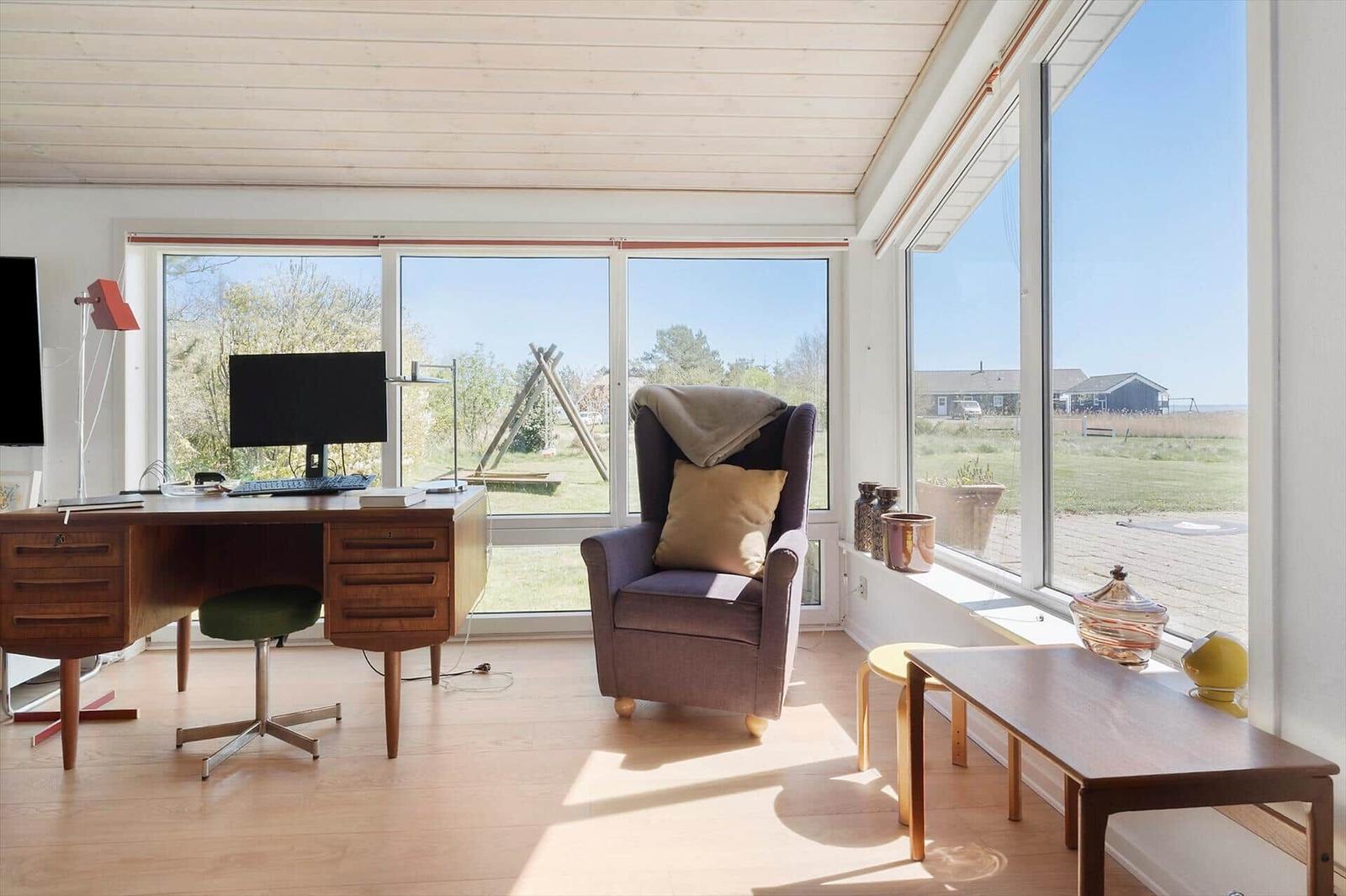 Ein Arbeitszimmer mit Schreibtisch, Stuhl und Sessel vor großen Fenstern.