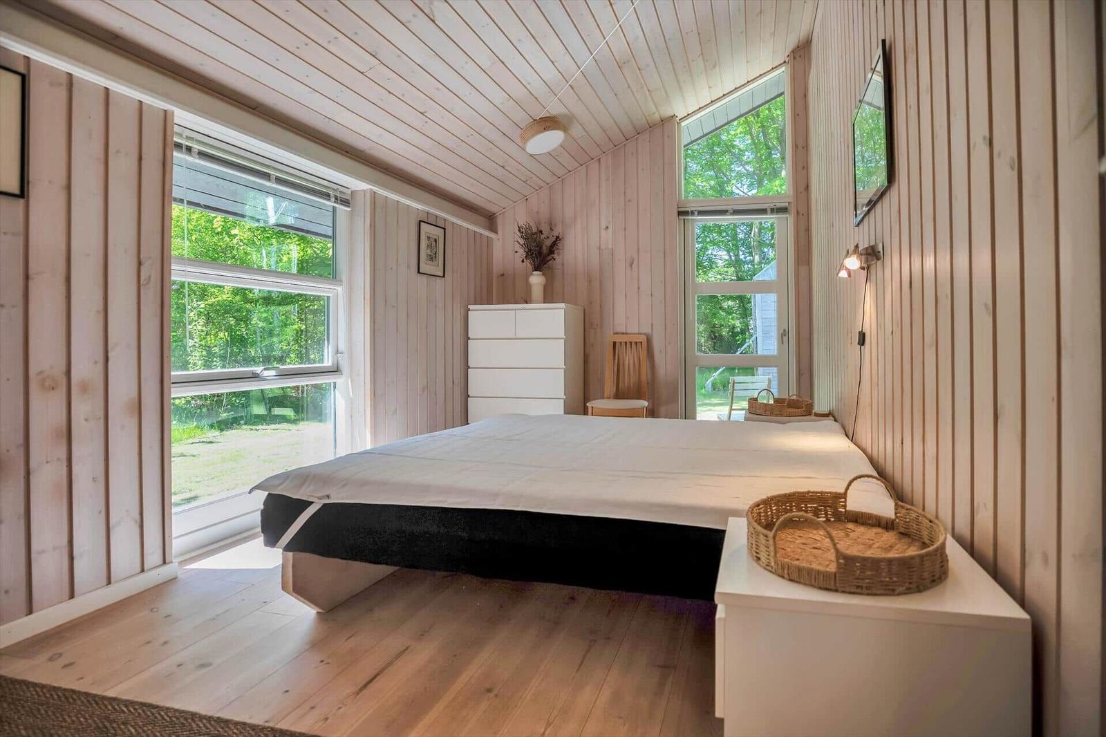 Schlafzimmer mit Holzwand, großem Bett und Fenstern zum Garten.