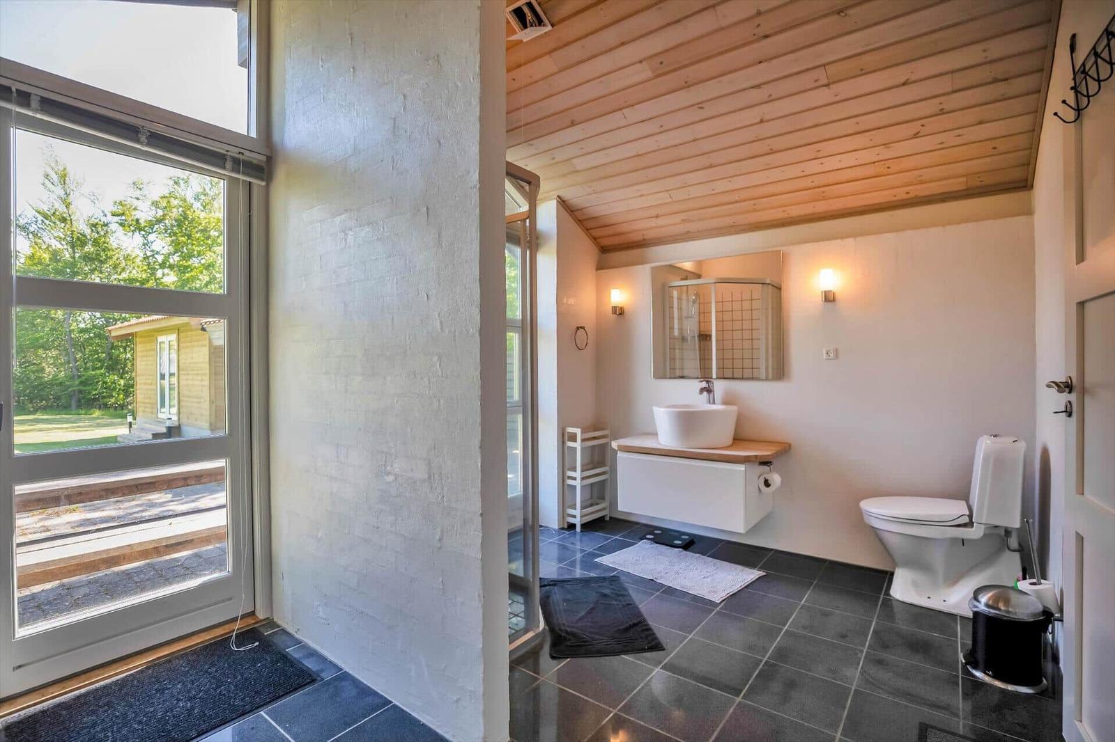 Badezimmer mit Toilette, Waschbecken und Dusche. Holzdecke und Fenster mit Blick auf Außenbereich.