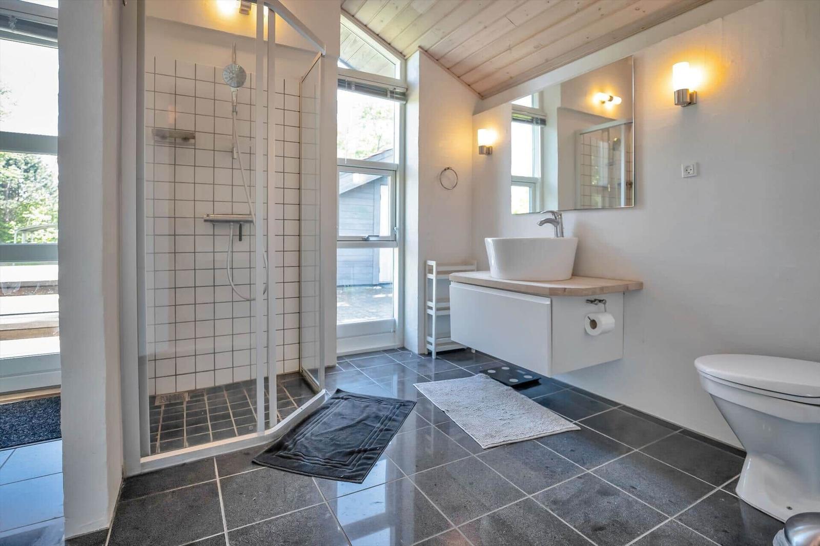 Badezimmer mit Dusche, Waschbecken und Toilette. Holzdecke und große Fenster.