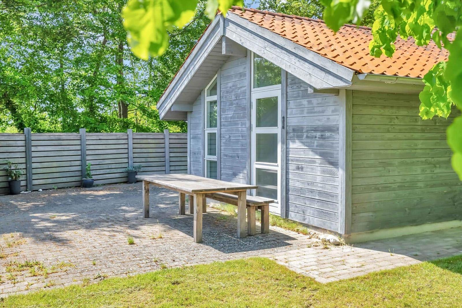 Holzhaus mit Terrasse, Tisch und Bänken im Garten. Umgeben von Grün und Zaun.