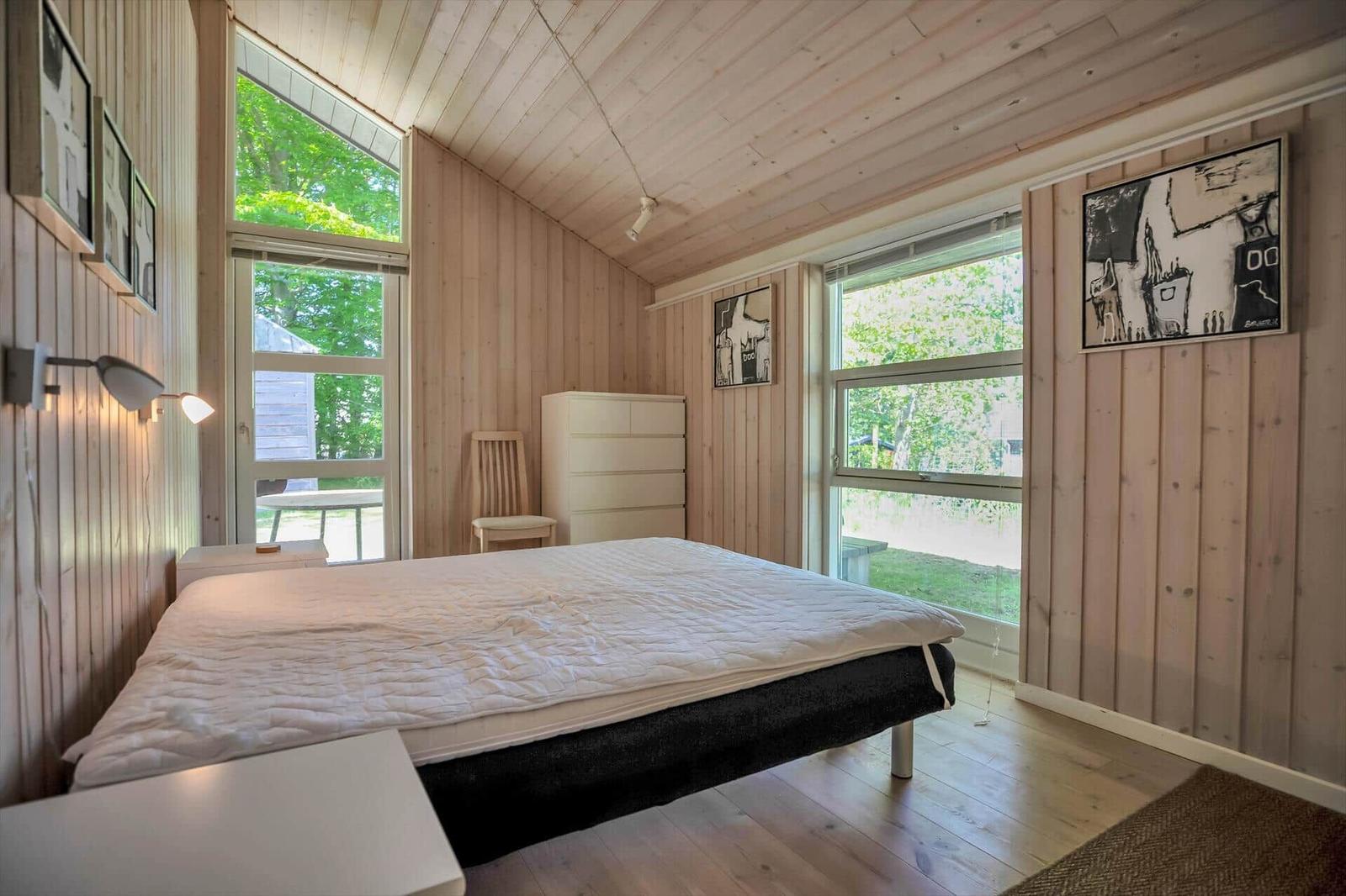 Schlafzimmer mit Holzwand, großem Bett und Fenstern zum Garten.