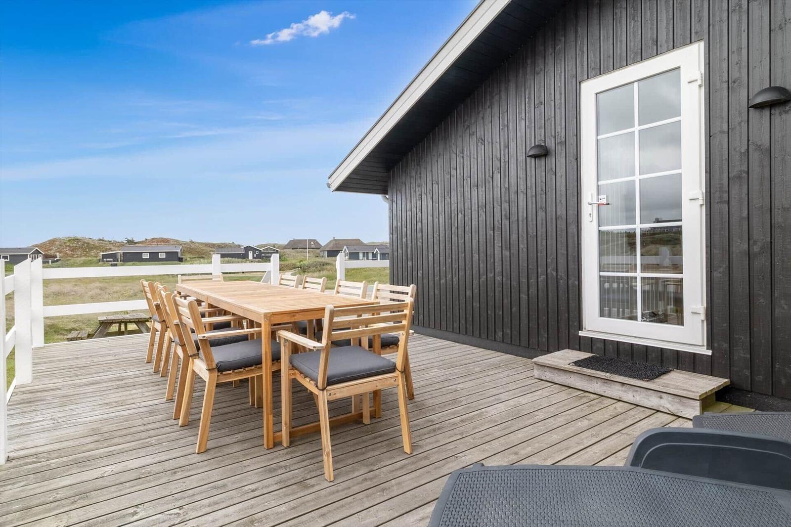 Terrasse mit Holztisch und Stühlen vor einem Haus mit dunkler Holzfassade und weißer Tür.