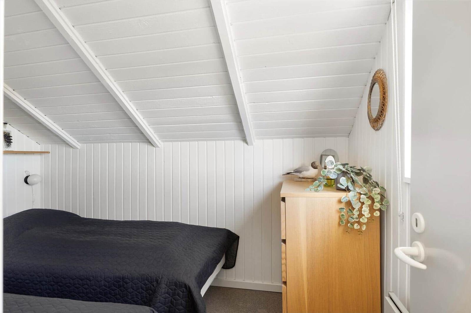 Schlafzimmer mit Dachschräge, weißer Wandverkleidung und Holzschrank. Auf Schrank: Vogel, Pflanze und Spiegel.