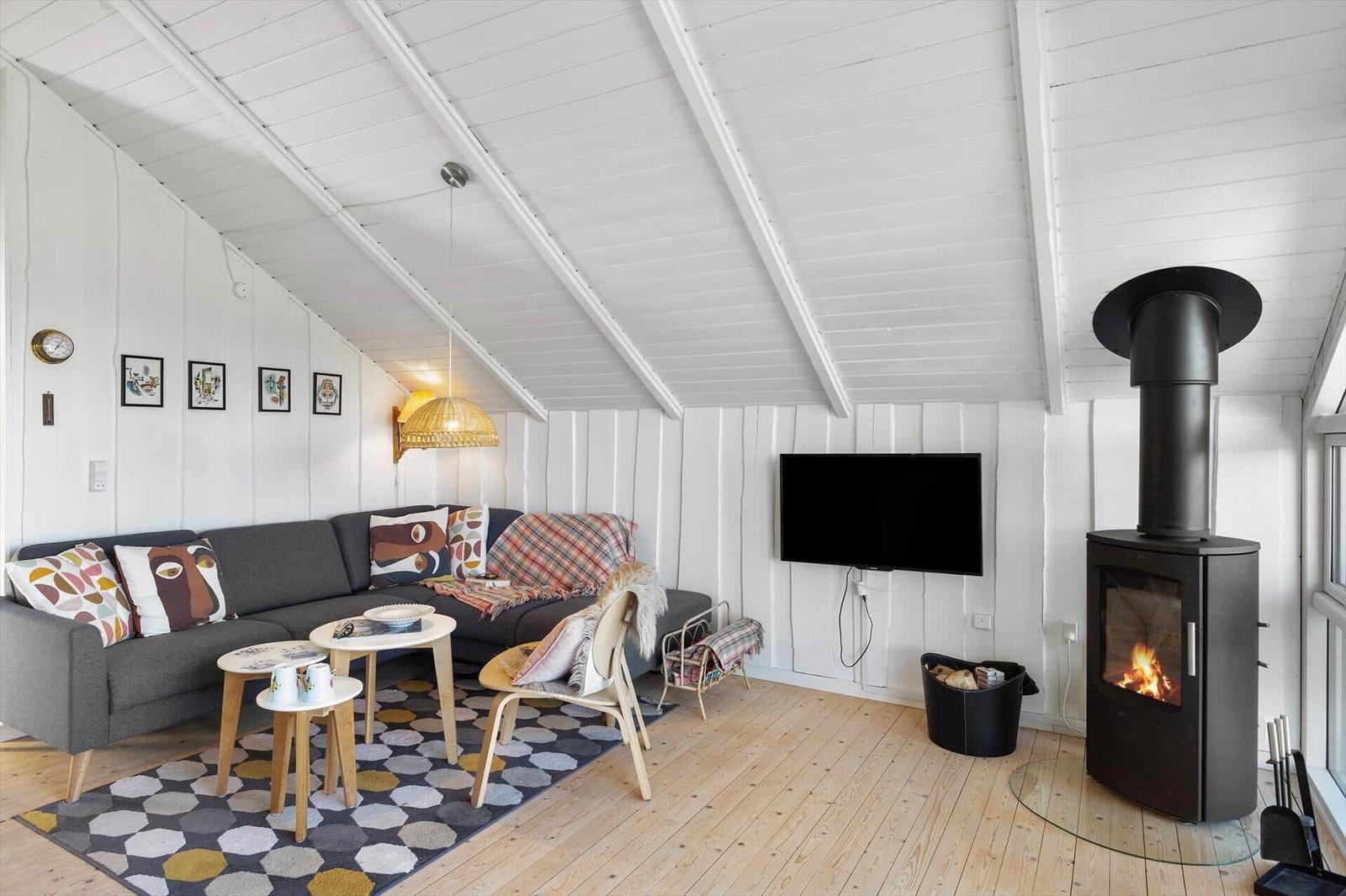 Wohnzimmer mit Sofa, Fernseher und Kaminofen. Holzboden und weiß verkleidete Wände.