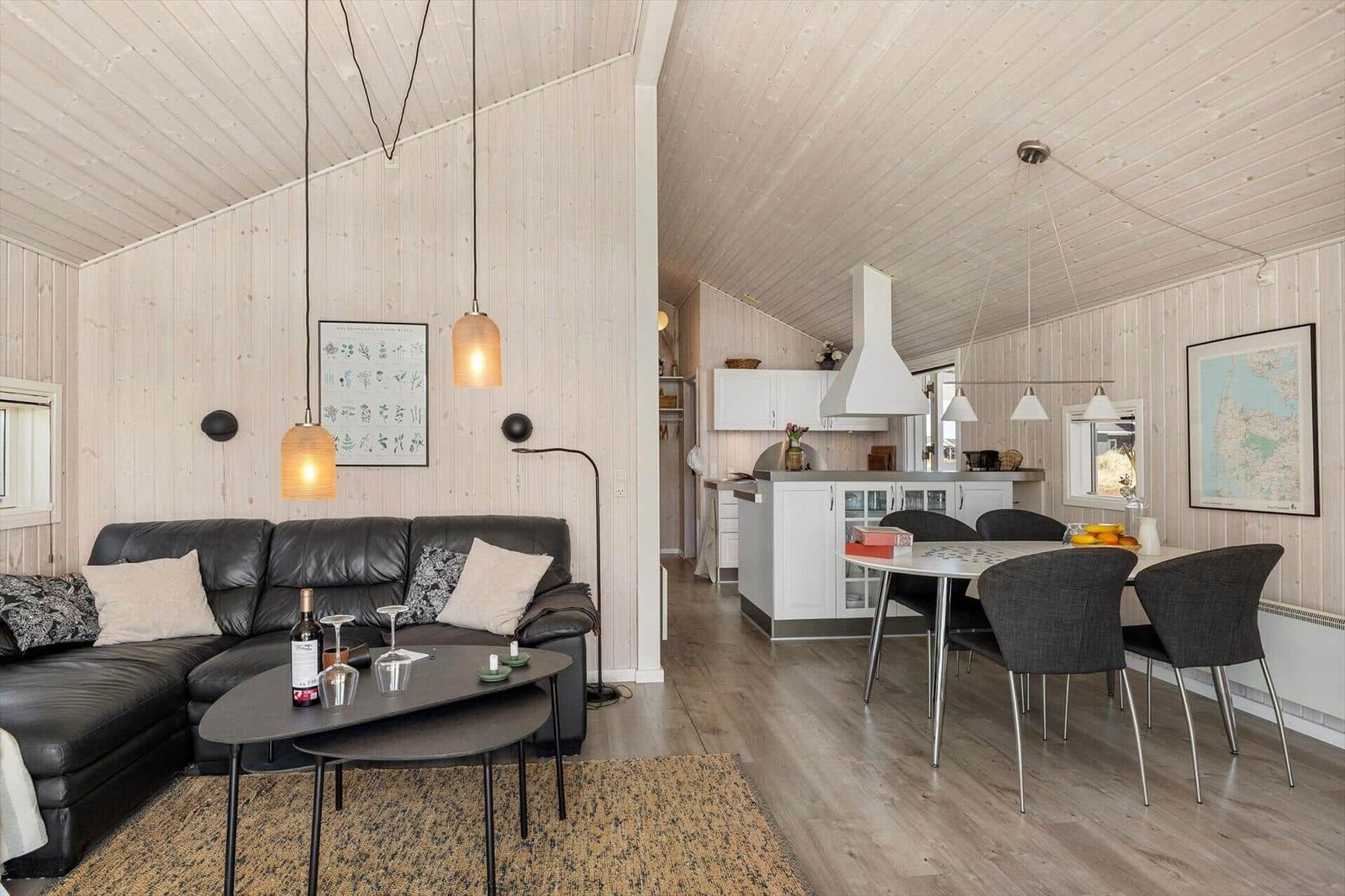 Wohn- und Essbereich mit Küche. Holzwände, schwarze Couch und Esstisch mit Stühlen.