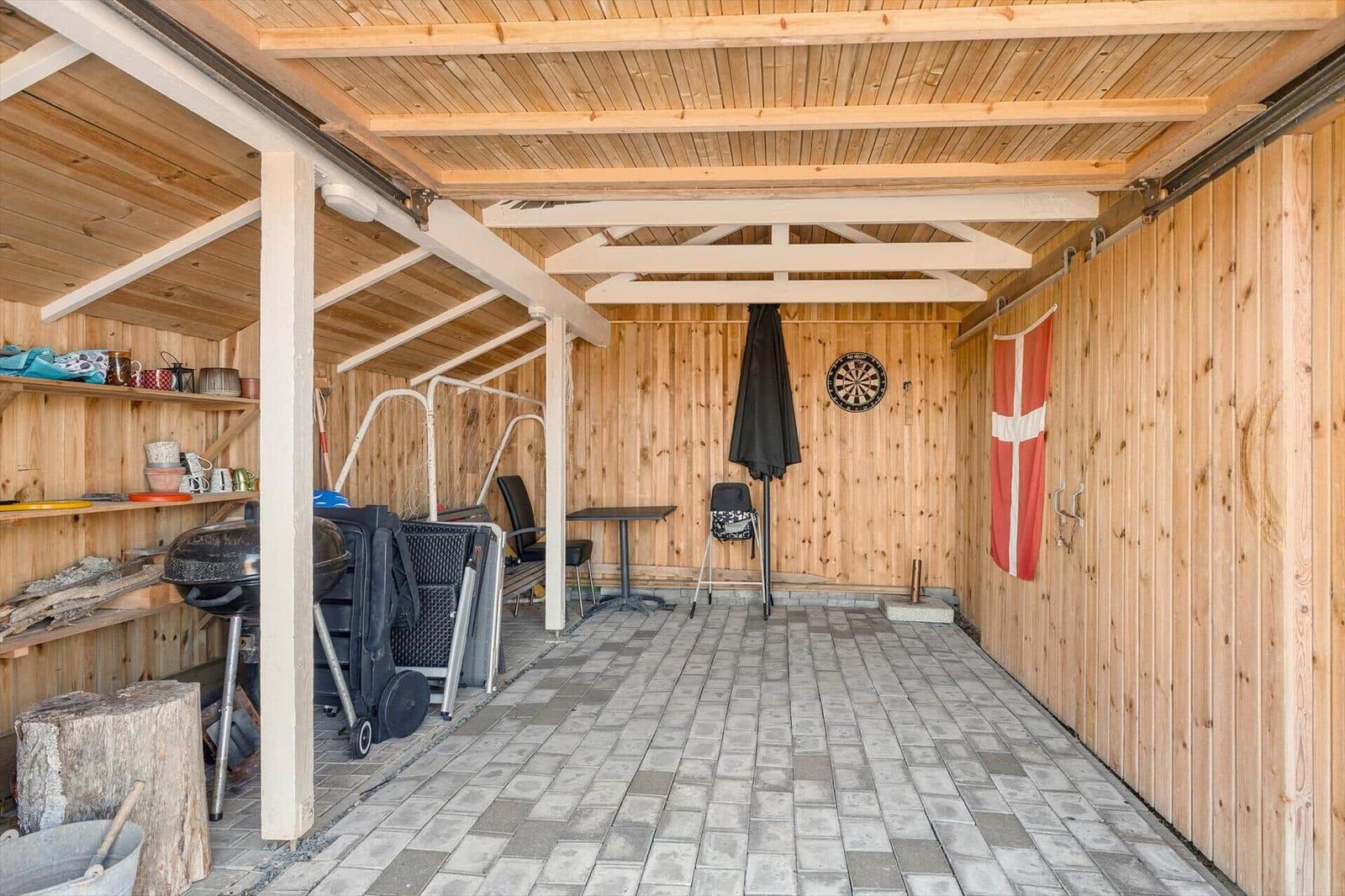 Holzüberdachter Bereich mit Grill, Tisch, Stühlen und Dartscheibe an der Wand.