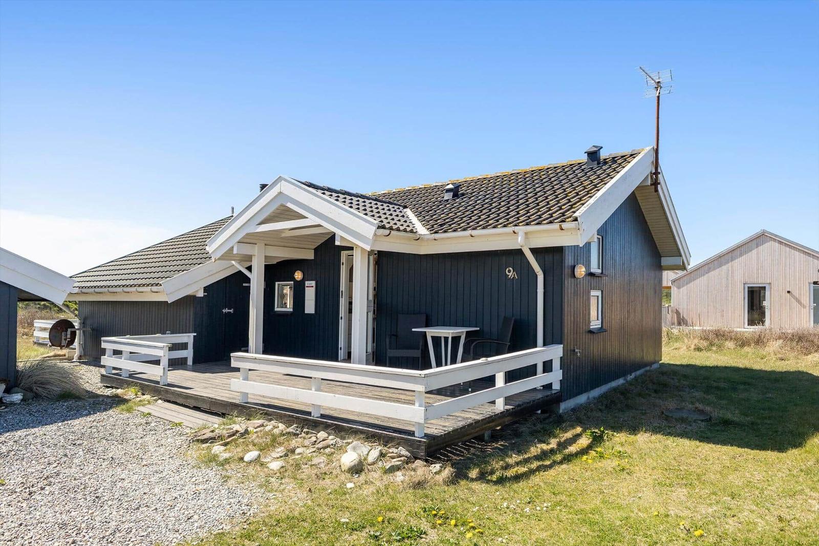 Ein dunkelblaues Ferienhaus mit weißem Balkon und Tisch. Hintergrund: Gras und klare Himmel.