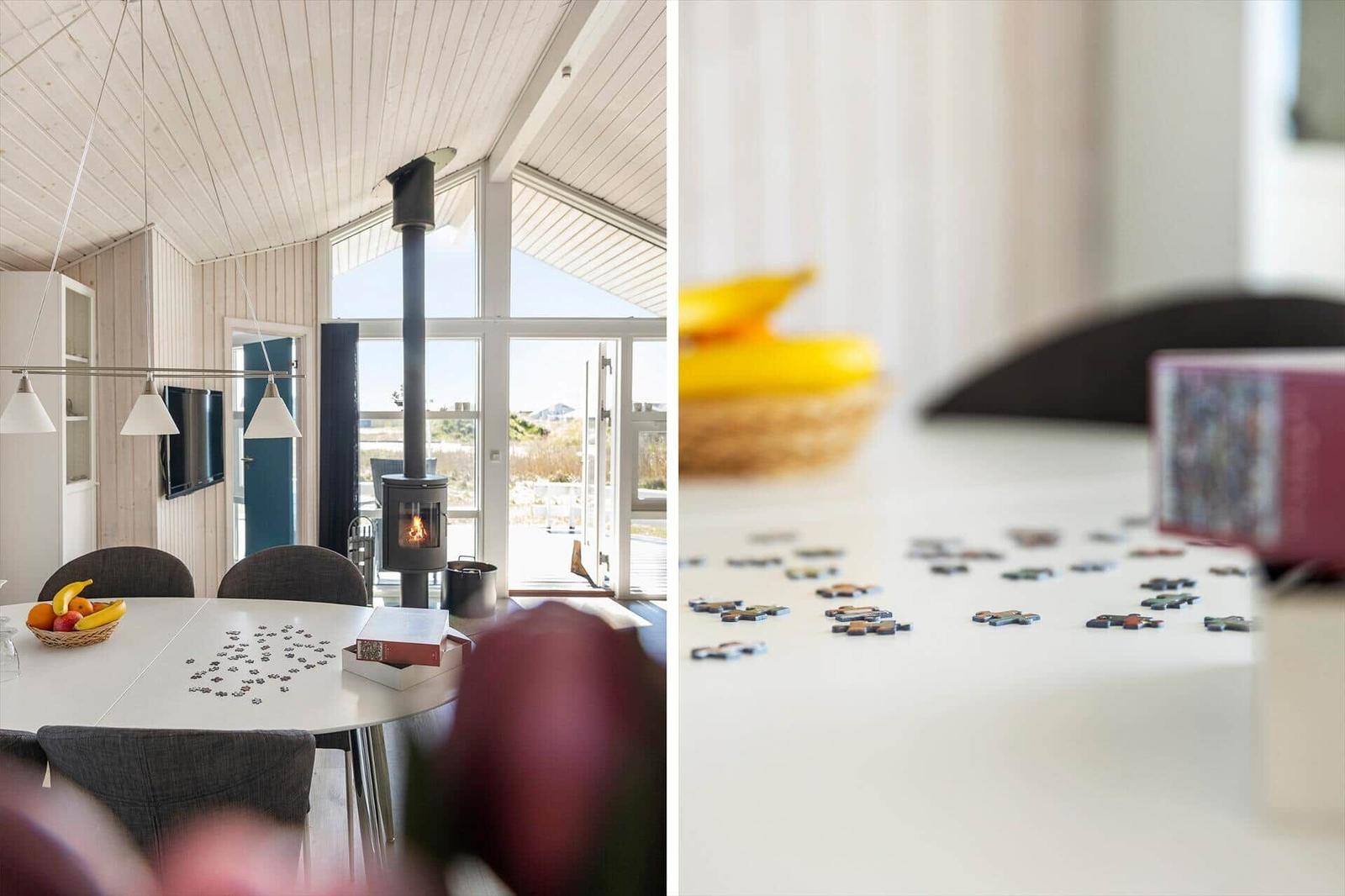 Esszimmer mit Esstisch, Puzzle und Holzofen. Große Fenster mit Blick auf Außenbereich.