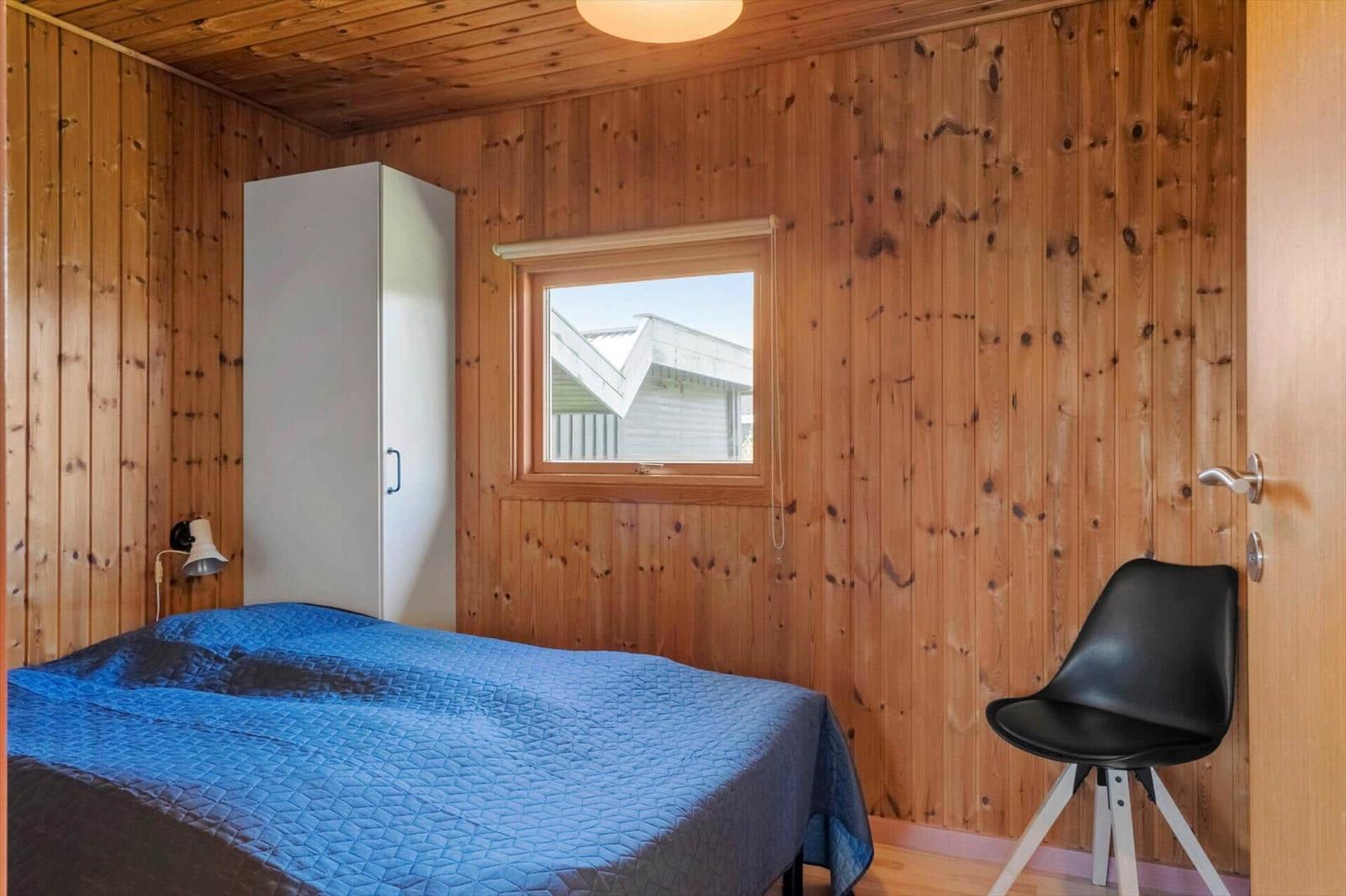 Schlafzimmer mit Holzwand, Bett mit blauer Bettdecke, Kleiderschrank und Stuhl.