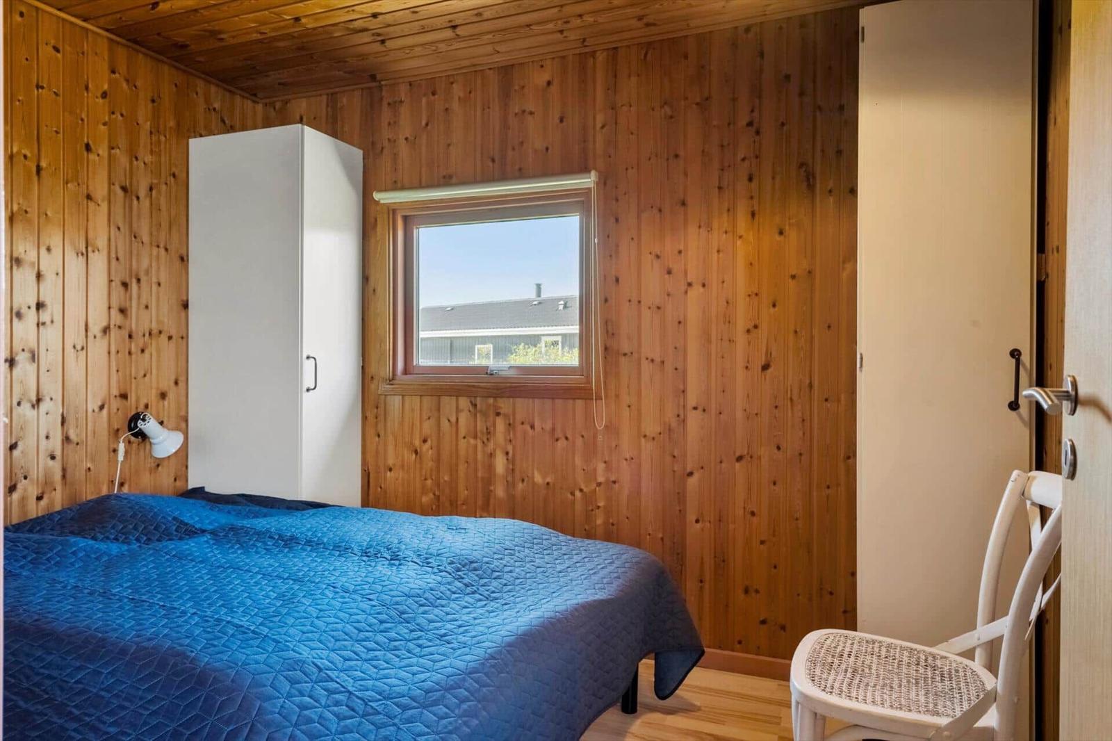 Schlafzimmer mit Holzwänden, blauem Bett und weißem Schrank. Fenster mit Blick auf Außenbereich.