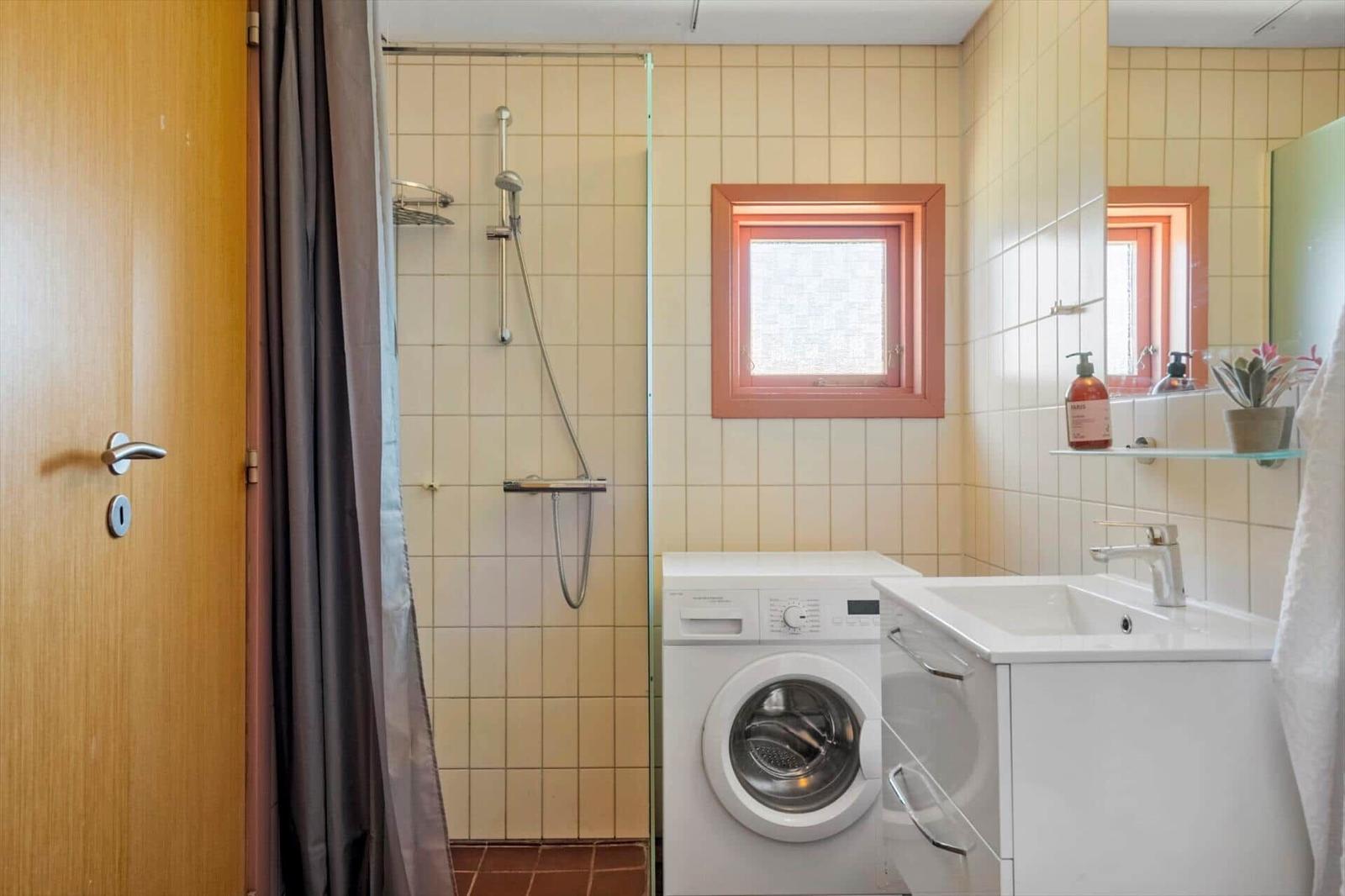 Badezimmer mit Dusche, Waschmaschine und Waschbecken. Fenster mit orangefarbenem Rahmen.