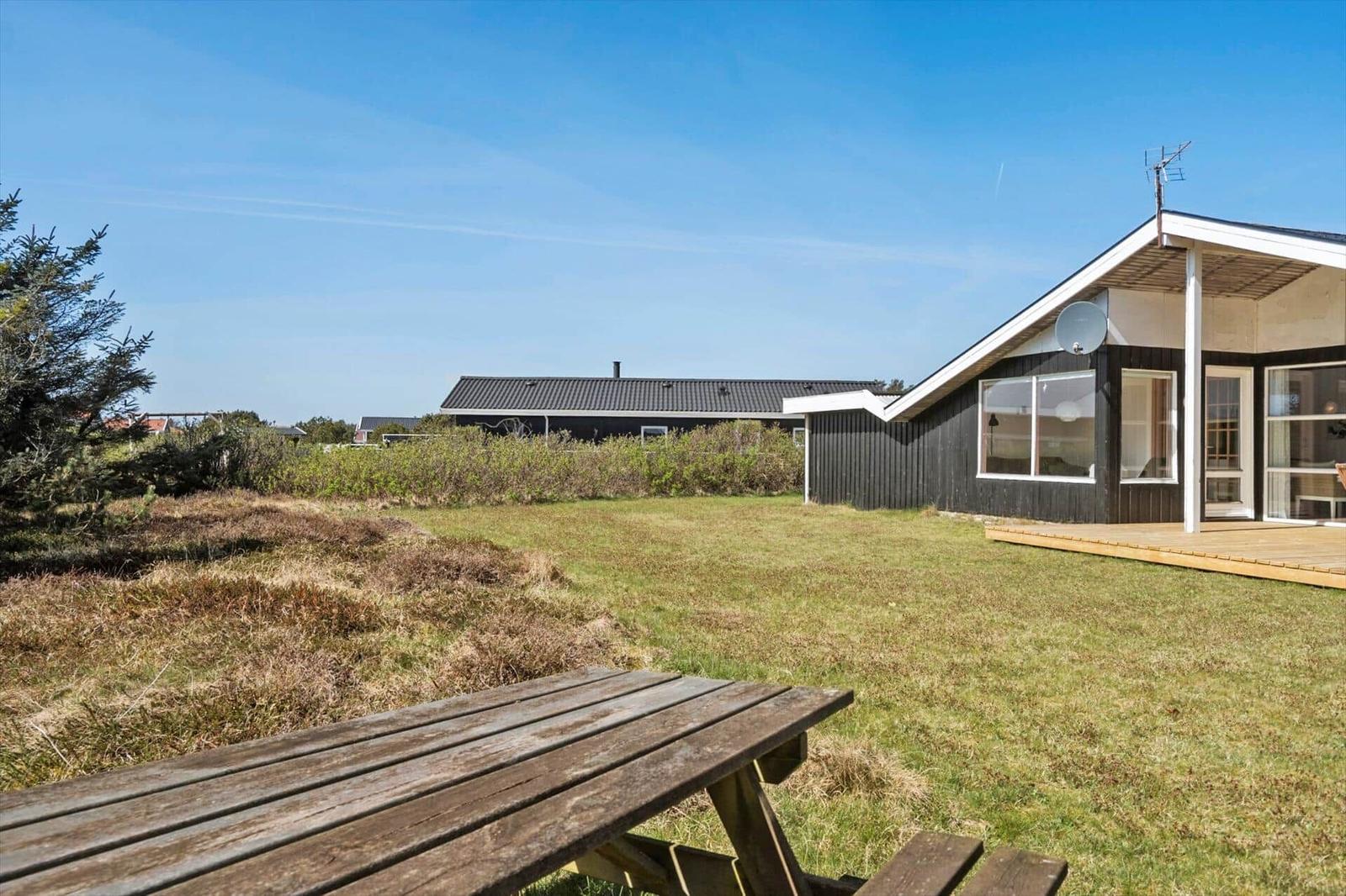 Haus mit Holzterrasse und Garten. Picknicktisch im Vordergrund. Himmel klar.