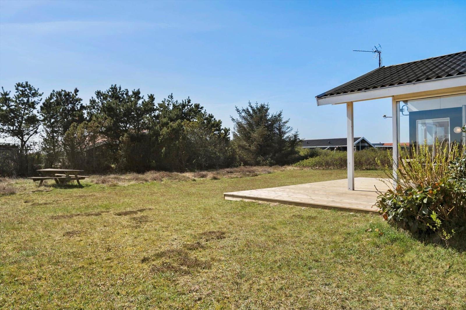 Haus mit Holzterrasse, Grasfläche und Picknicktisch. Hintergrund: Bäume und klarer Himmel.