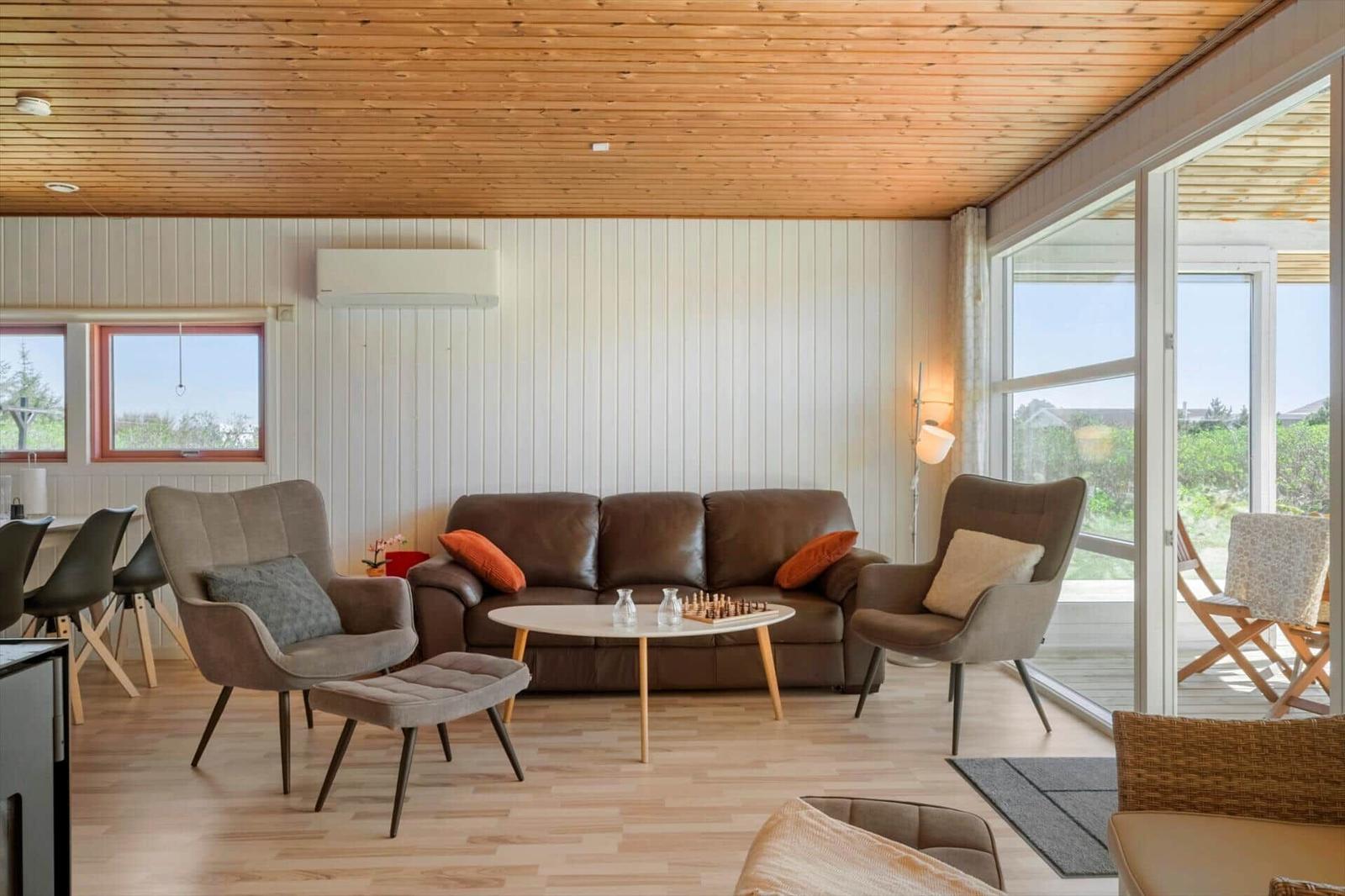 Wohnzimmer mit Sofa, zwei Sesseln und Tisch. Holzdecke und große Fenster.