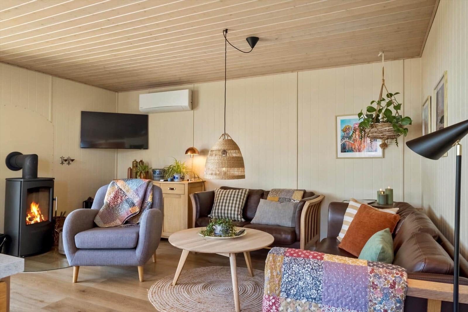 Wohnzimmer mit Kamin, Sofa und Sessel. Holzdecke, Pendelleuchte und Pflanzen.