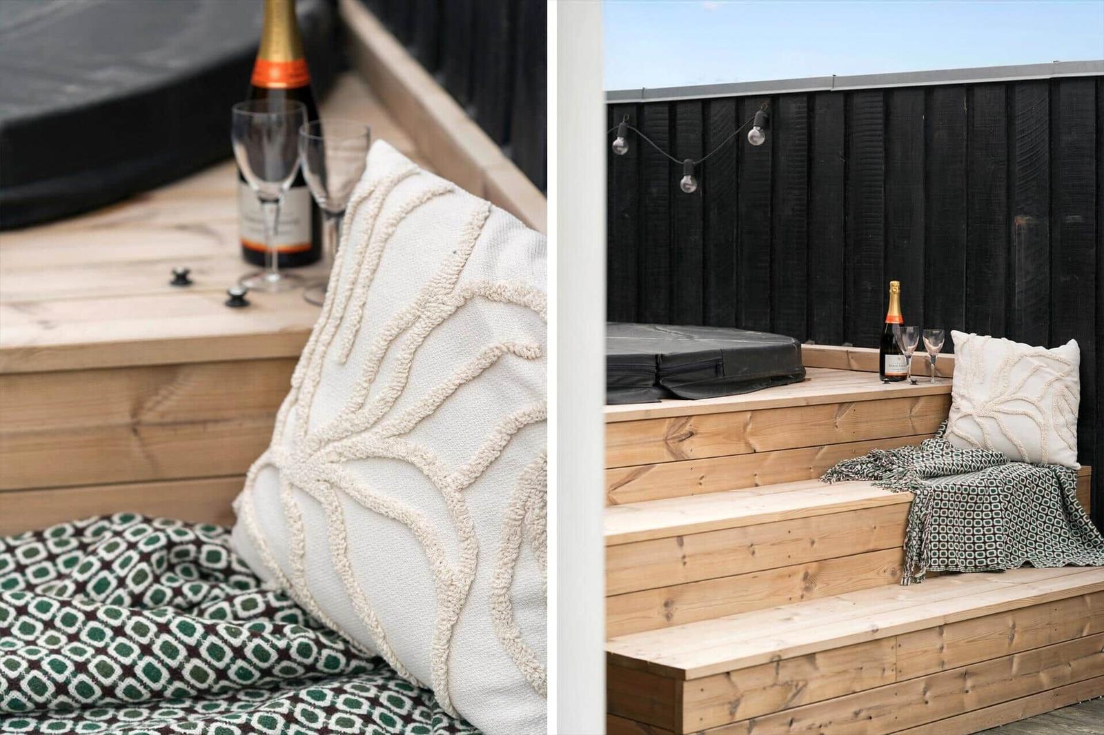 Terrasse mit Holzbank, Kissen und Champagner. Hintergrund: schwarze Holzwand und Lichterkette.