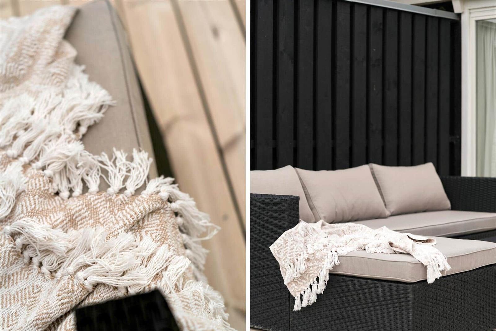 Außenliegende Sofa mit Kissen und Decke auf Terrasse vor schwarzer Holzwand.
