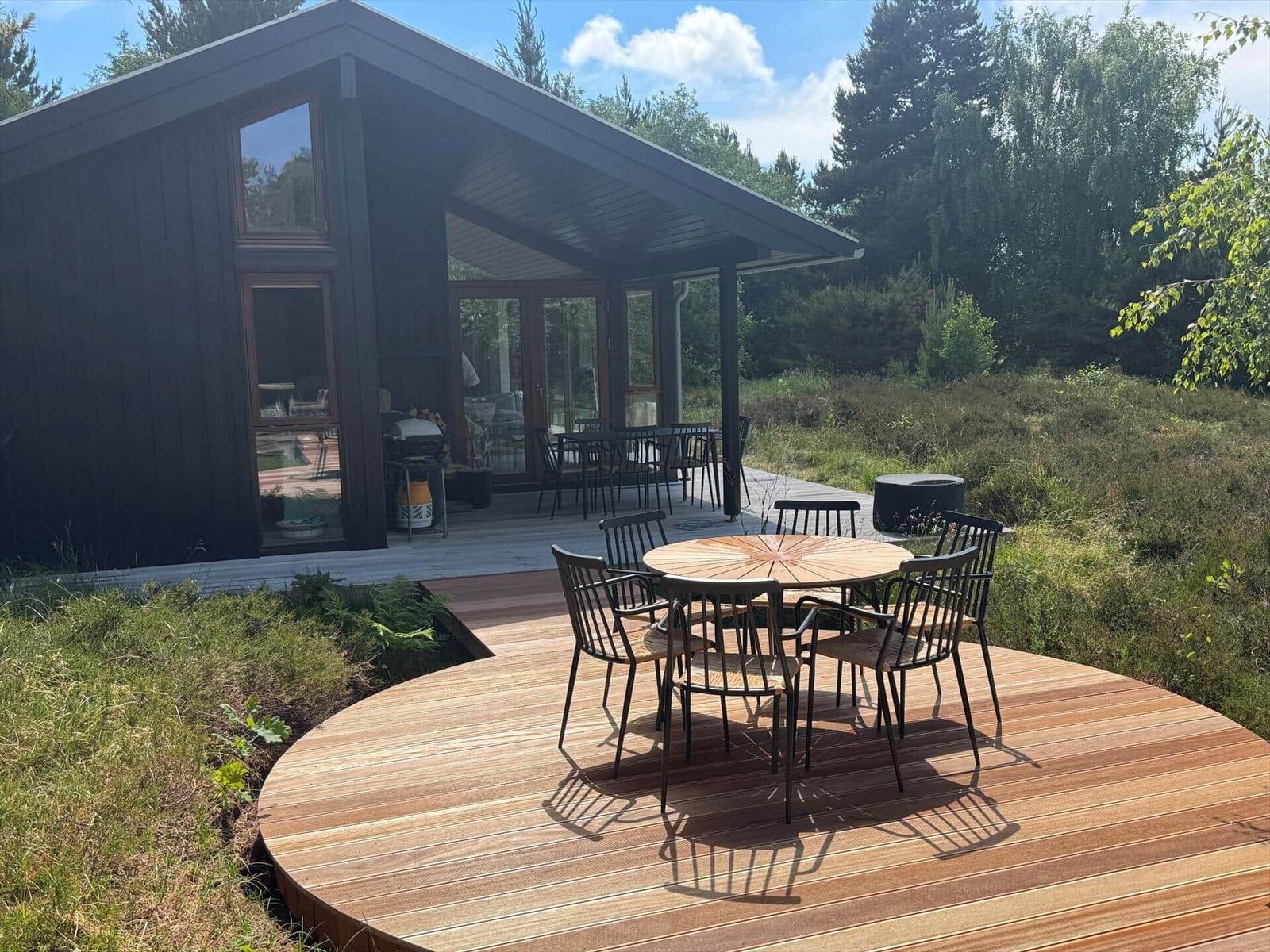 Moderne Holzhütte mit Terrasse, runder Tisch und Stühlen im Grünen.