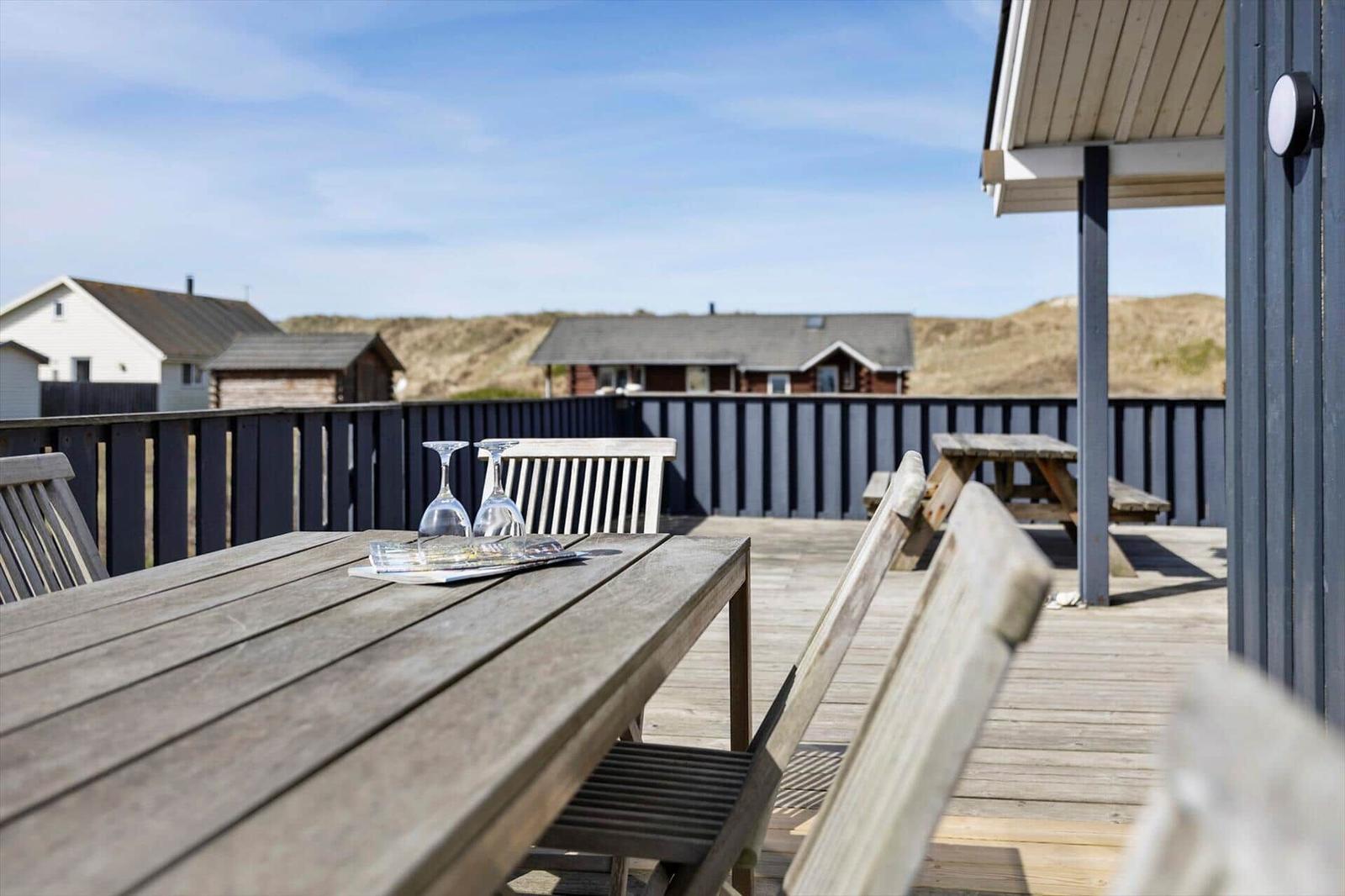 Terrasse mit Holztisch und Stühlen, Blick auf Strandhäuser und Dünen.