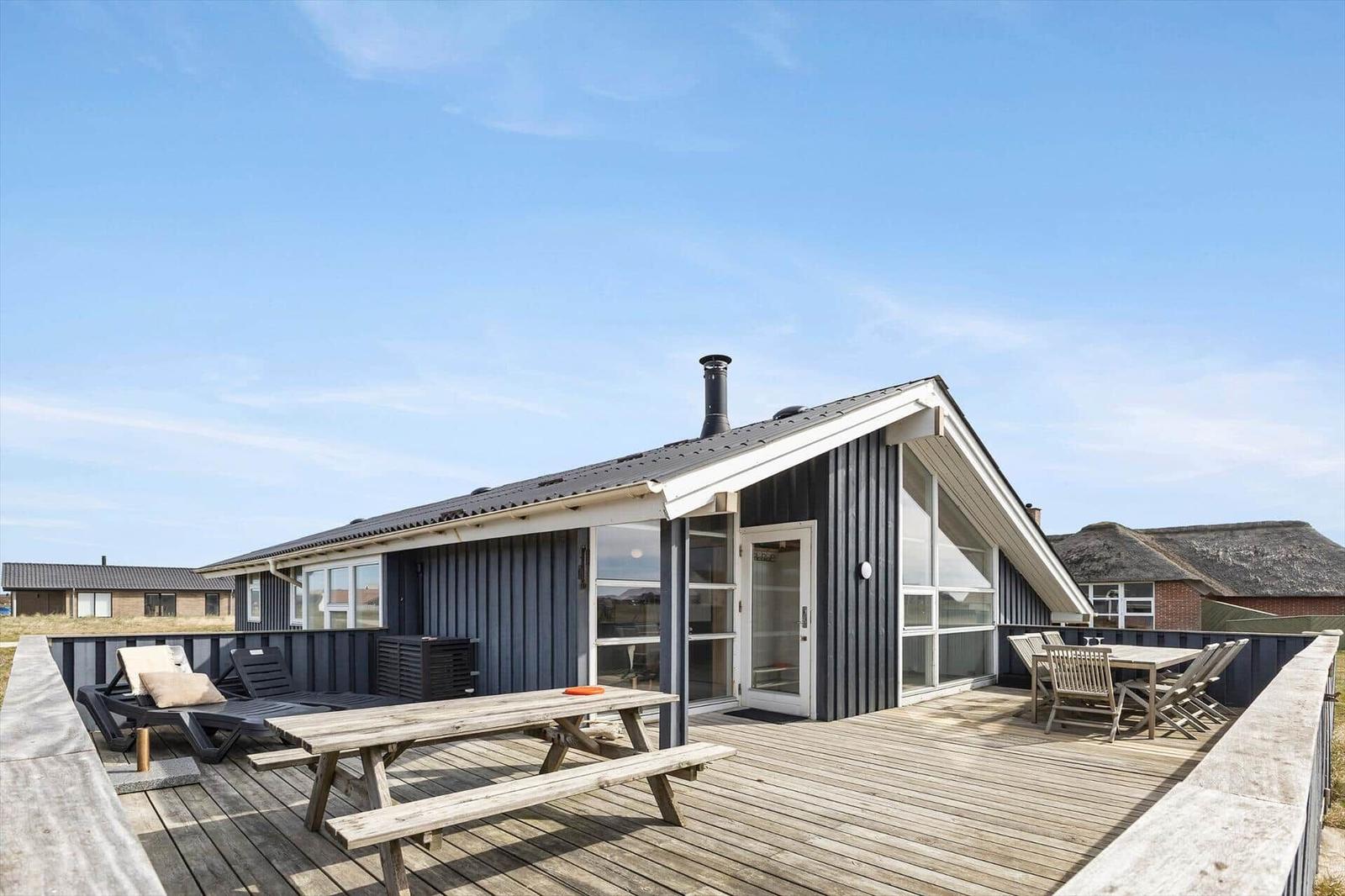 Moderne Villa mit großem Holzdeck, Picknicktisch und Liegestühlen unter blauem Himmel.