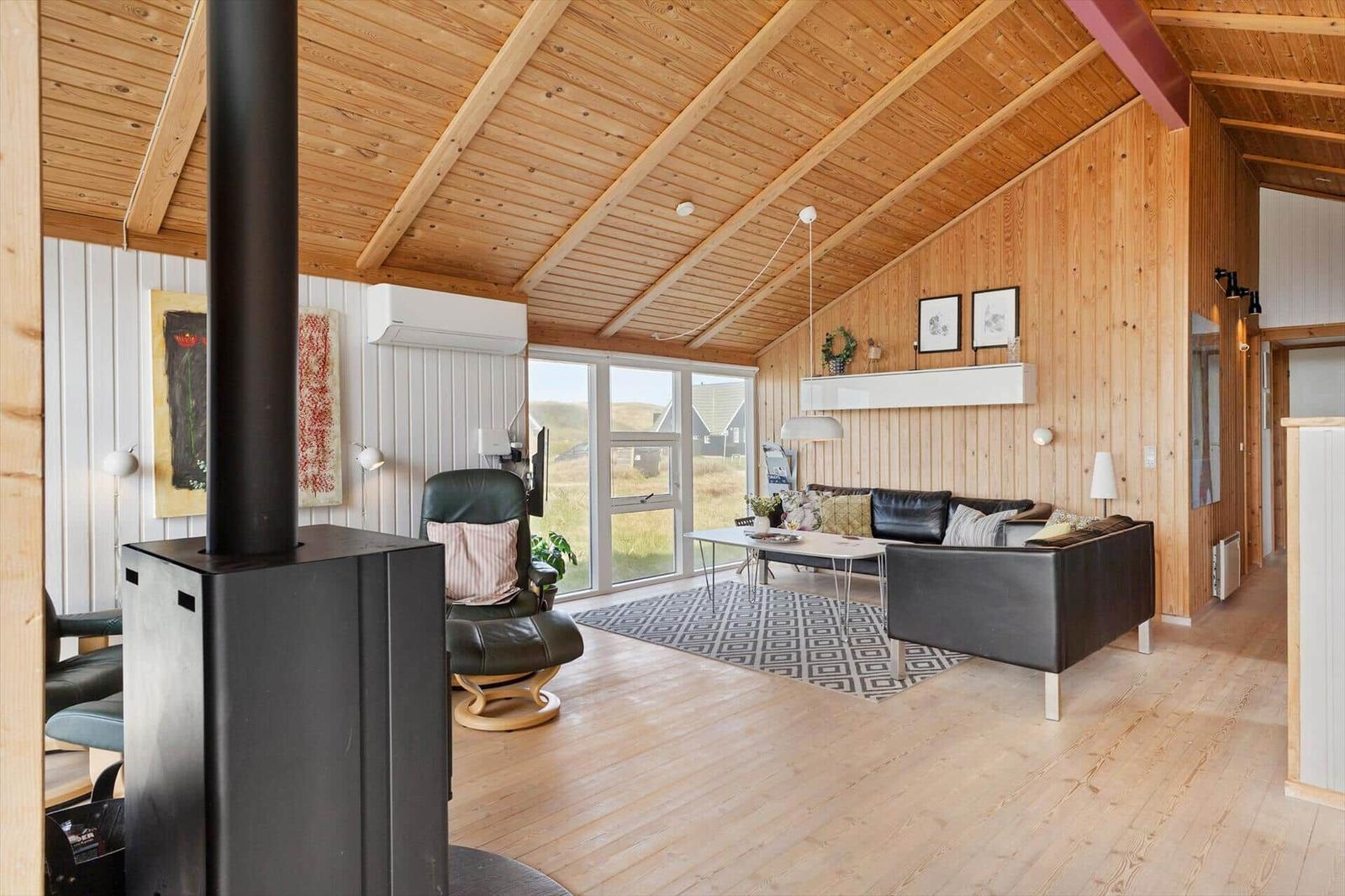 Wohnzimmer mit Holzdecke, Sofa, Sessel und Kaminofen. Große Fenster mit Blick auf die Landschaft.