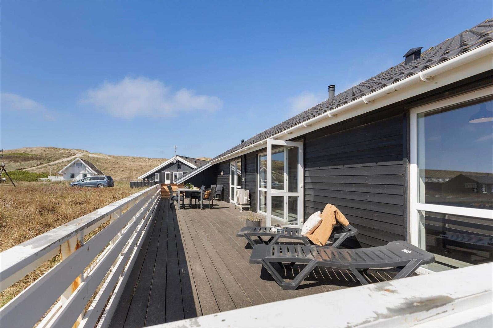 Terrasse mit Liegen und Tisch. Schwarzes Haus mit weißem Geländer. Blick auf Dünen und Himmel.