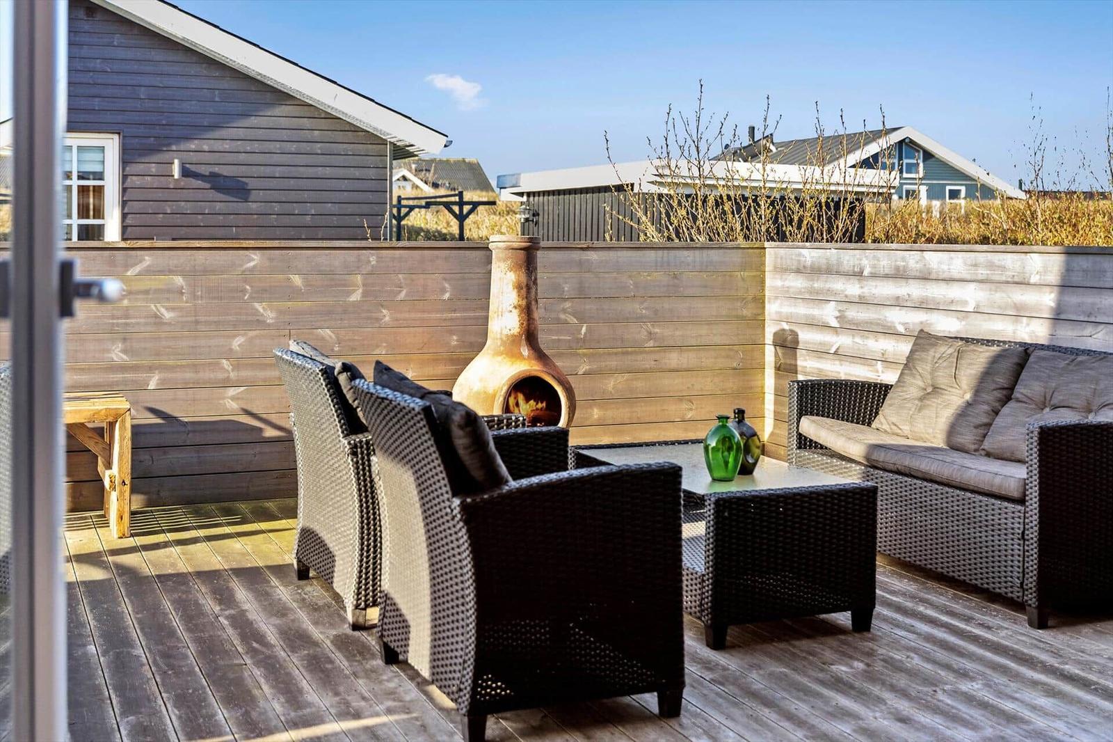 Terrasse mit Sitzgruppe, Feuerkamin und Holzgeländer. Hintergrund: Nachbarhäuser und trockener Busch.