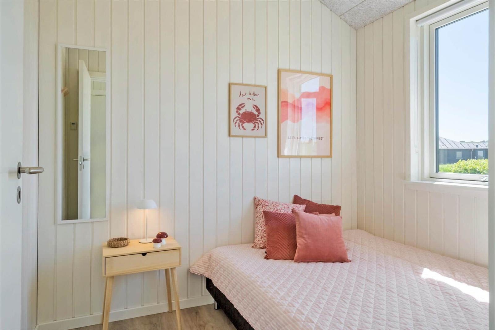 Schlafzimmer mit Bett, Nachttisch und Fenster. Wände sind weiß verkleidet.