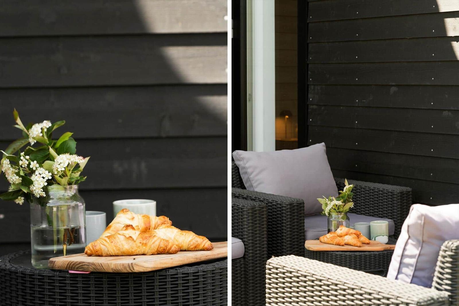 Außentisch mit Croissants, Blumen und Tassen vor schwarzem Holzhaus.