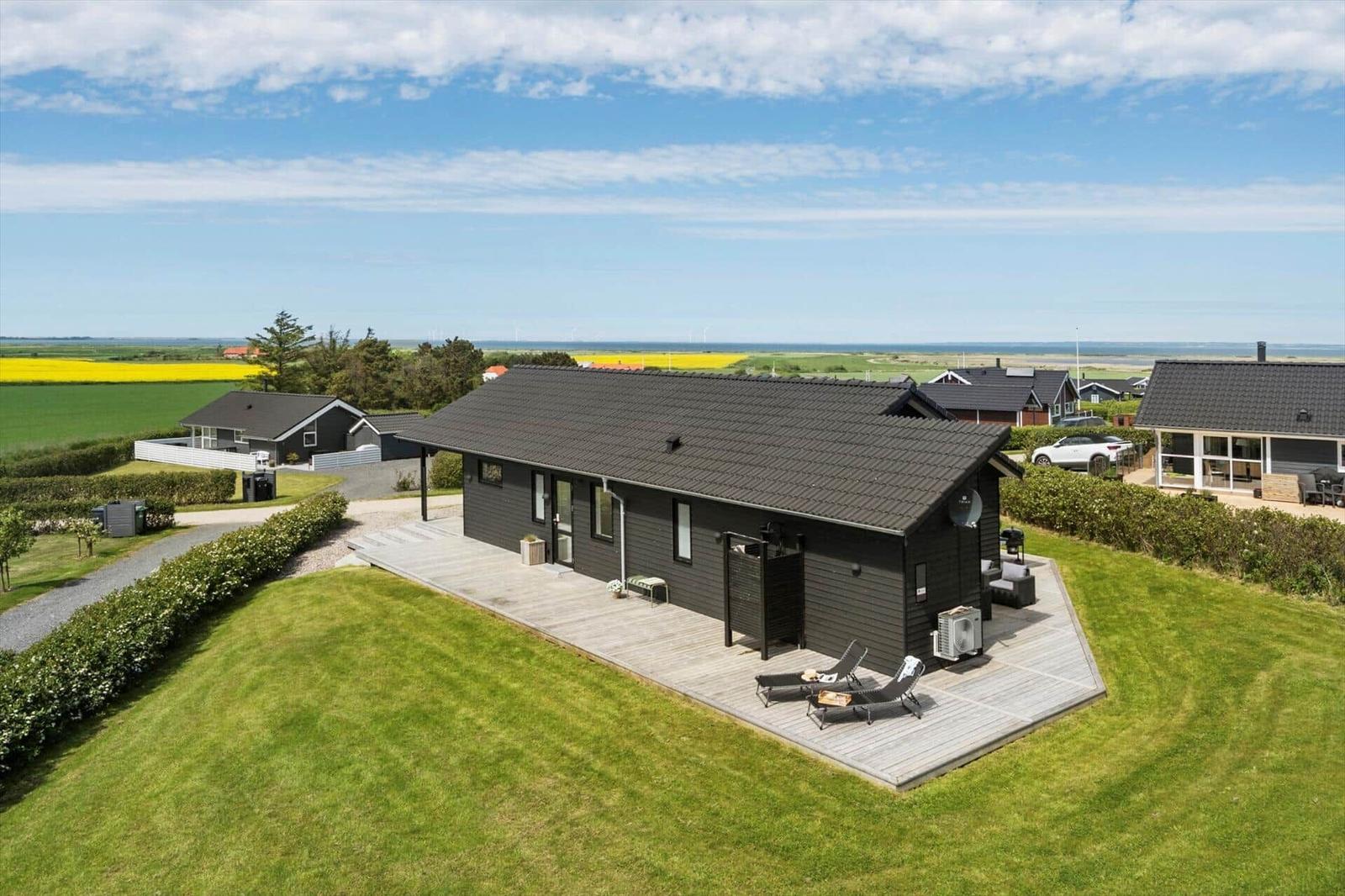 Ein modernes Haus mit Terrasse und Garten. Im Hintergrund sind Felder und das Meer zu sehen.