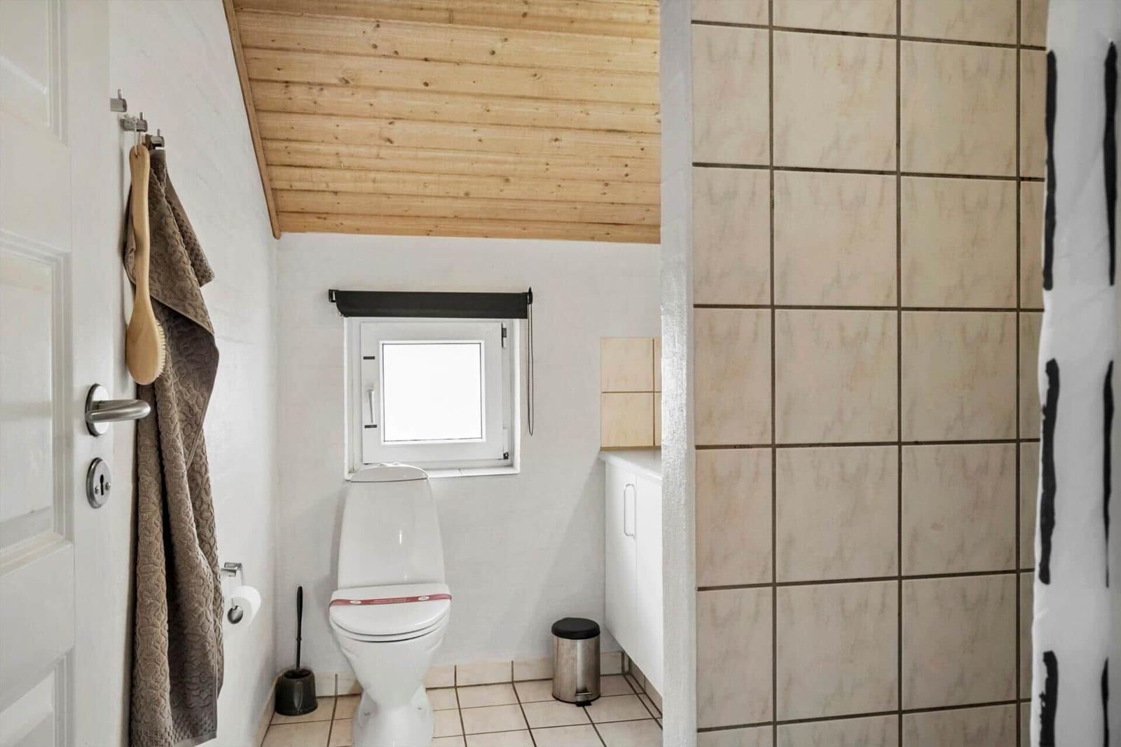 Badezimmer mit Toilette, Dusche und kleinem Fenster. Holzdecke und Fliesenwand.