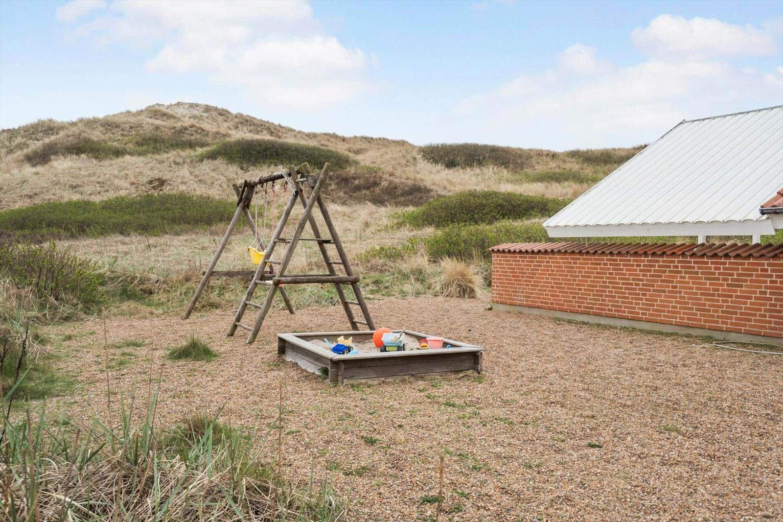 Hinterhof mit Sandkasten und Schaukel, umgeben von Dünen und Steinmauer.