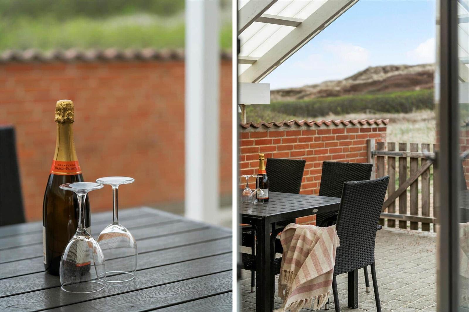 Terrasse mit Tisch, Stühlen und Champagner. Hintergrund: Ziegelmauer und Dünen.
