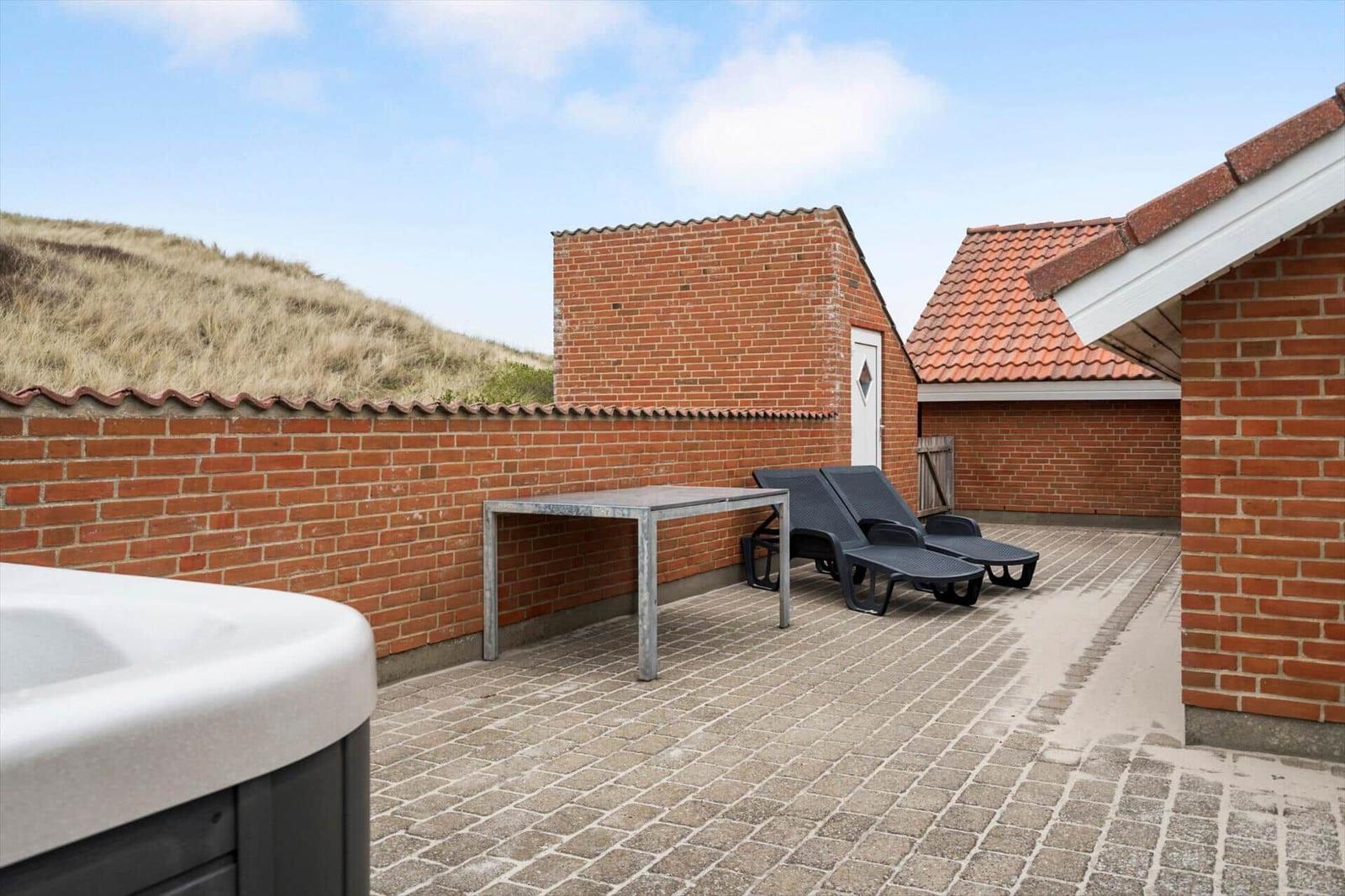 Terrasse mit Liegestühlen, Tisch und Whirlpool. Hintergrund: Ziegelmauer und Dünen.