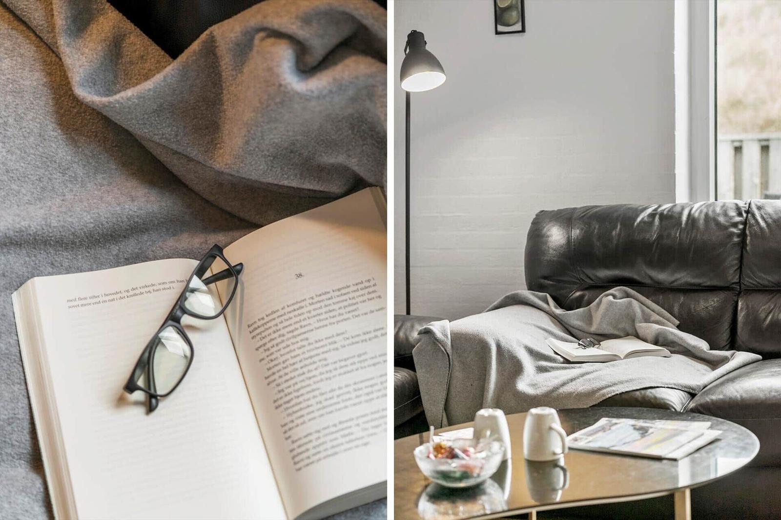 Ein Buch mit Brille liegt auf einer grauen Decke. Ein schwarzer Ledersofa mit Decke und Tisch.