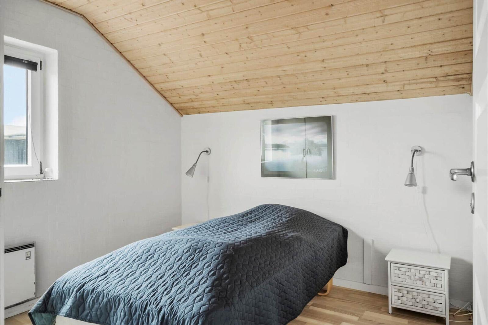 Schlafzimmer mit Holzdecke, Bett mit blauer Bettdecke, Nachttisch und Wandlampen.