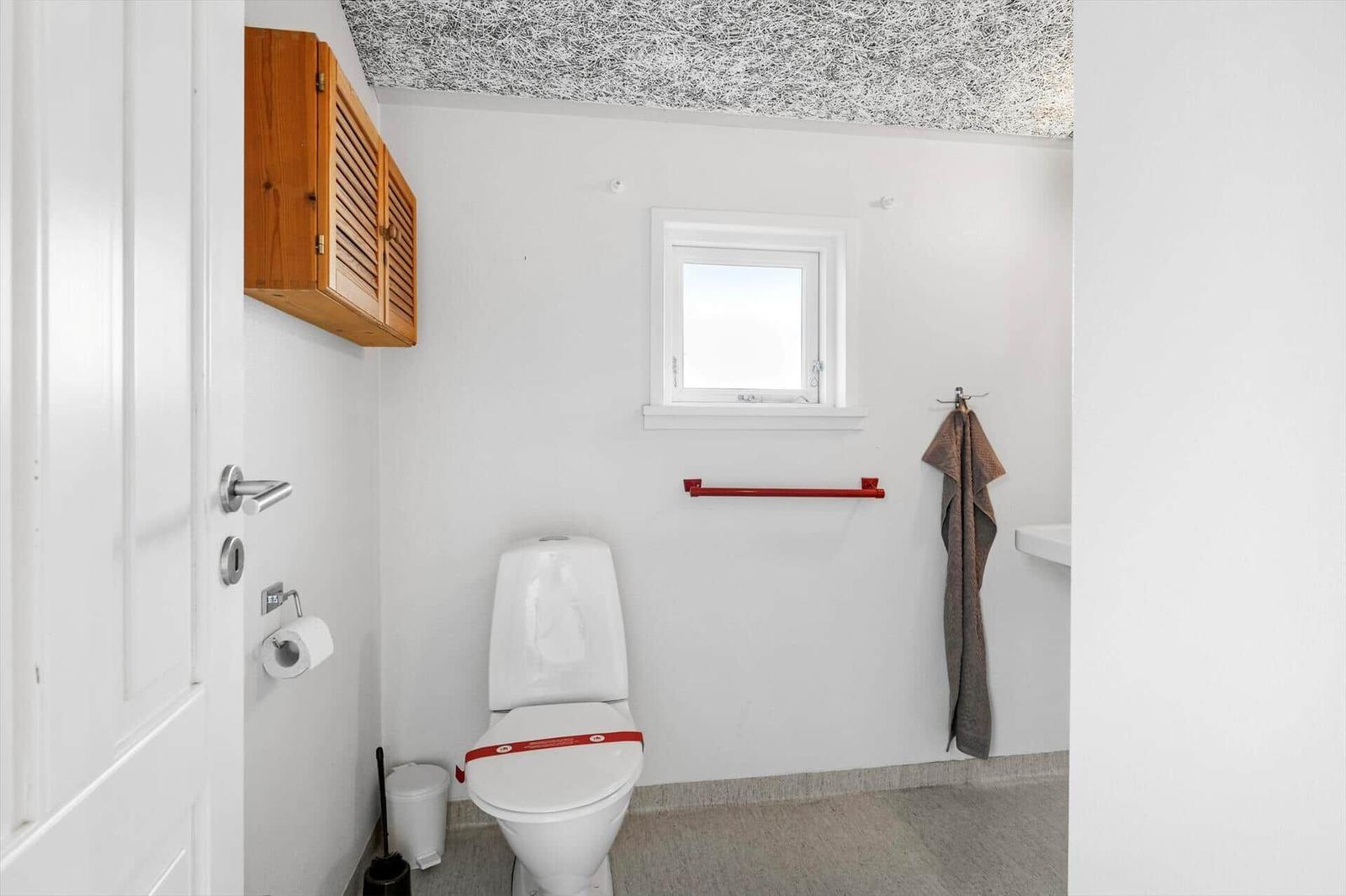 Badezimmer mit Toilette, Fenster, Handtuch und Holzschränkchen an der Wand.