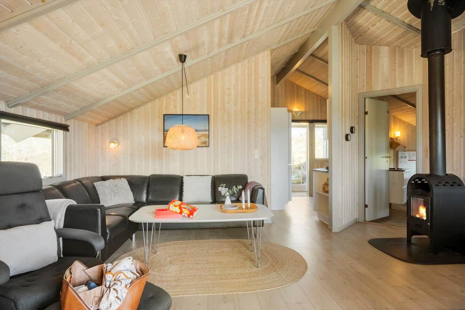 Wohnzimmer mit Sofa, Tisch und Kaminofen. Holzwände und -decke. Küche im Hintergrund.