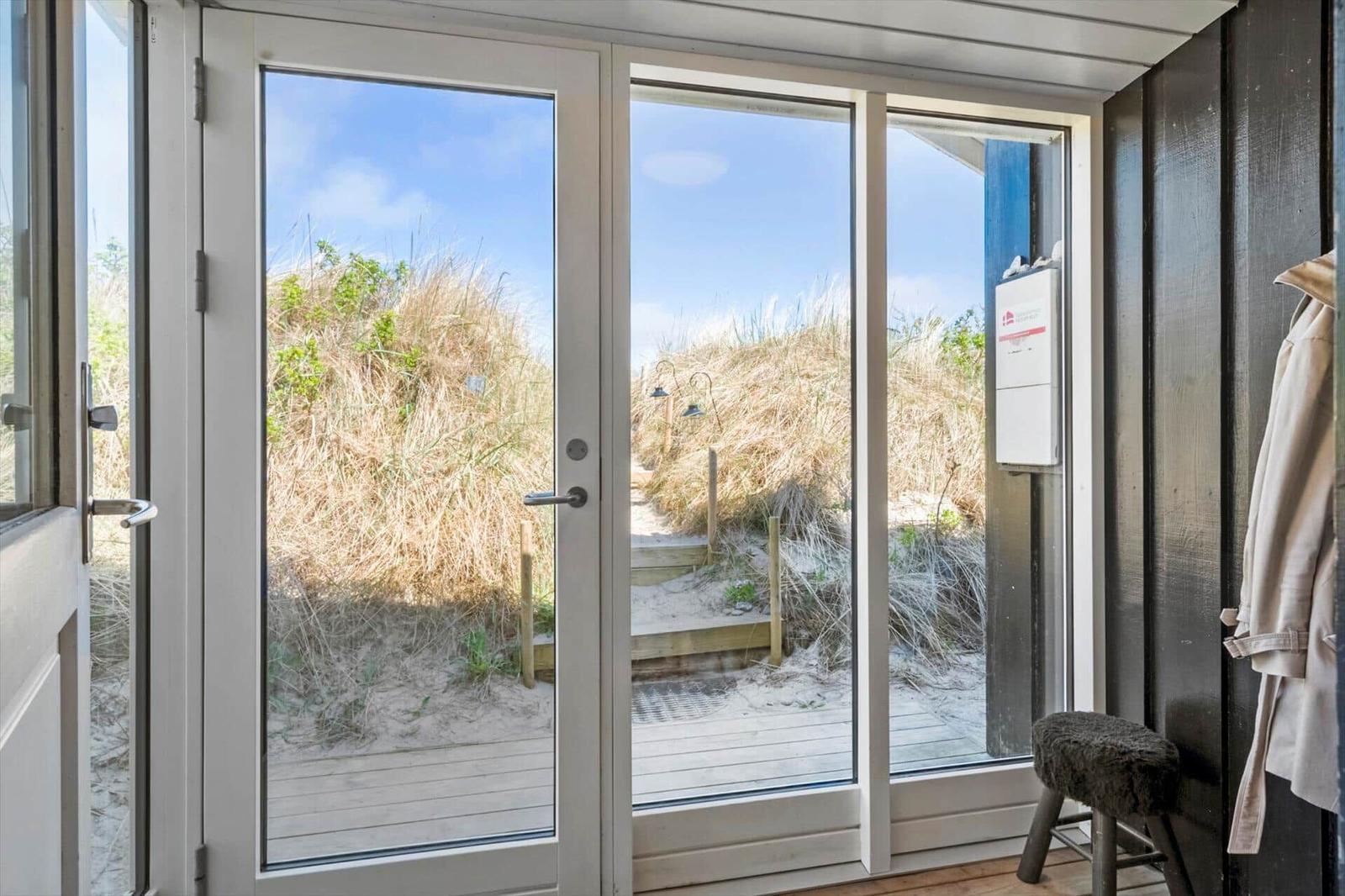 Durch die Glastüren blickt man auf einen Sandstrand mit Dünen und einem Holzweg.