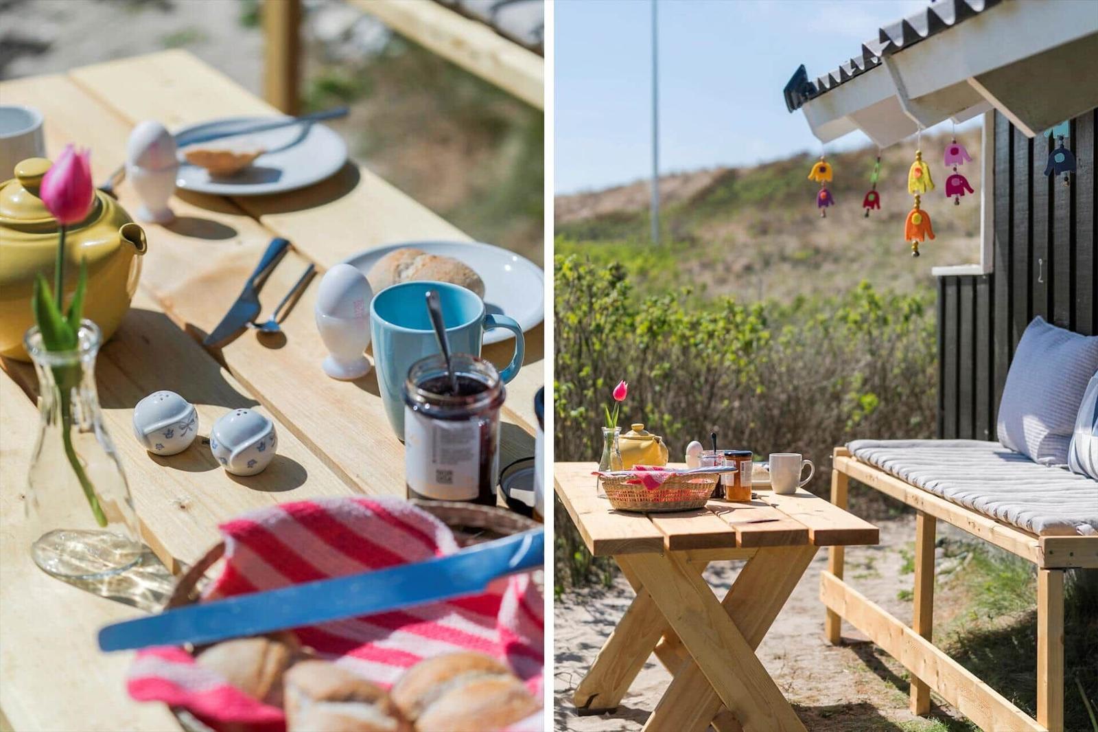 Holztisch mit Frühstück im Freien. Hintergrund: Strandhütte mit Sitzbank und Windspiel.