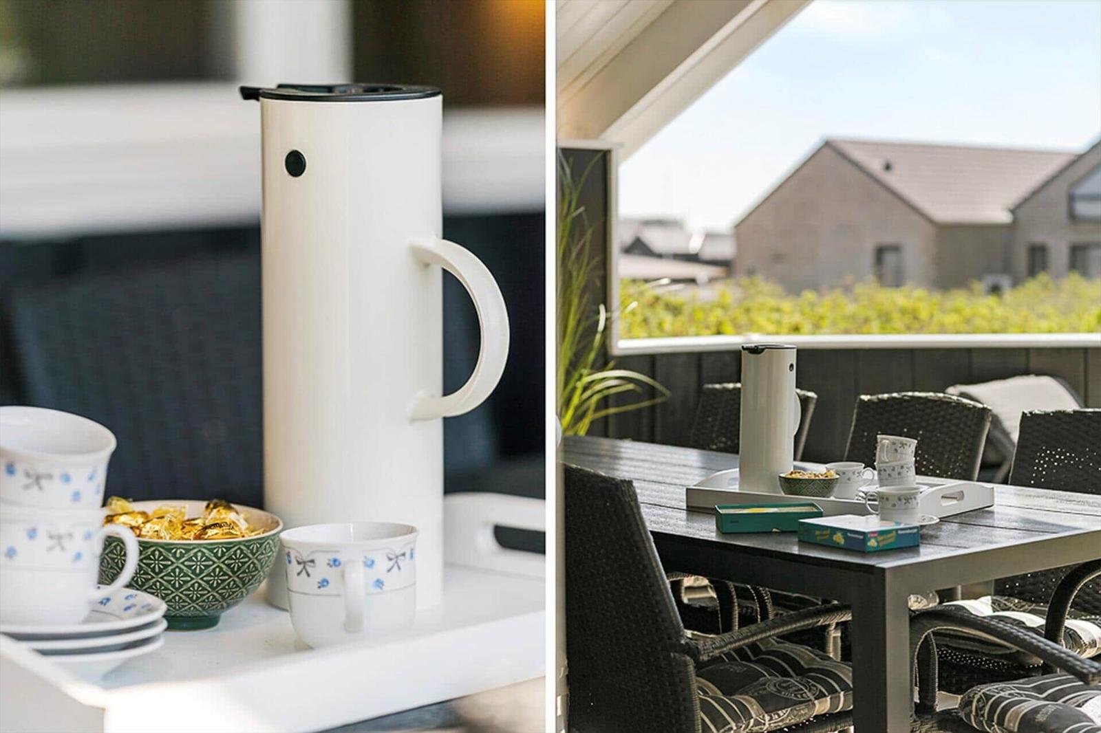 Terrasse mit Tisch, Stühlen und Kaffeekanne. Blick auf grüne Pflanzen und Gebäude.