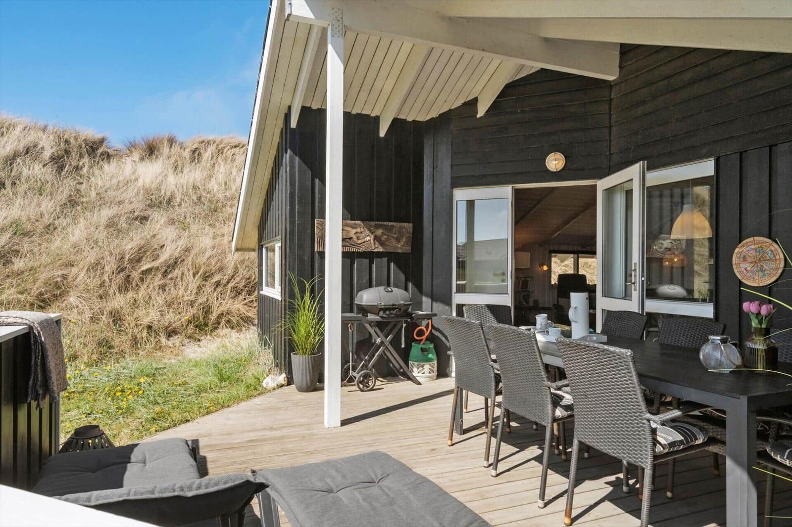 Terrasse mit Grill, Tisch und Stühlen. Hintergrund: Dünen und Haus mit offenen Türen.