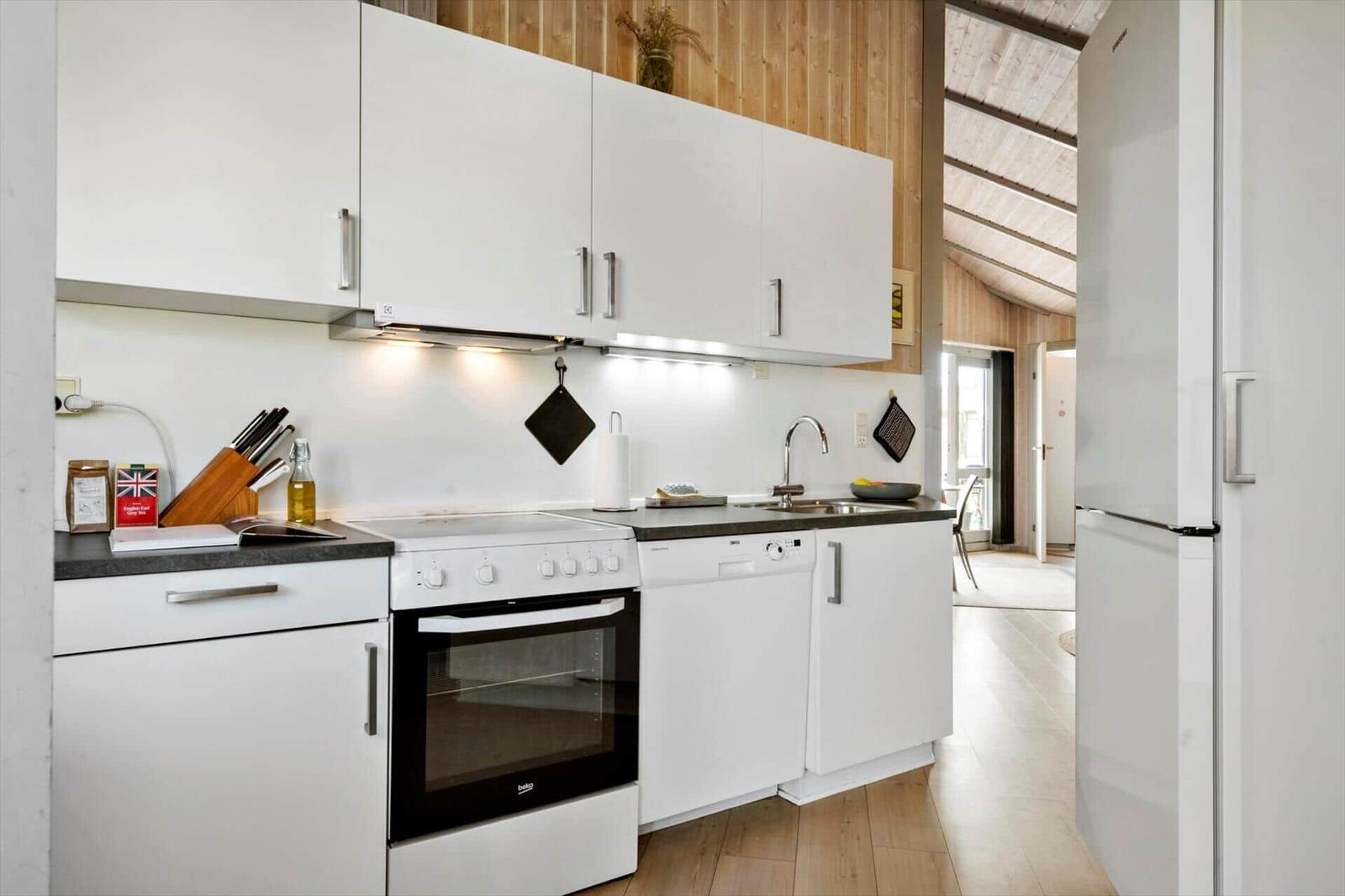 Küche mit weißen Schränken, Edelstahlspüle und Kühlschrank. Holzwand und Lichtdurchlass sichtbar.