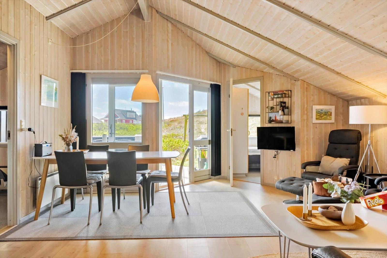 Wohnraum mit Essbereich, TV und Sitzgelegenheit. Holzwände und große Fenster.