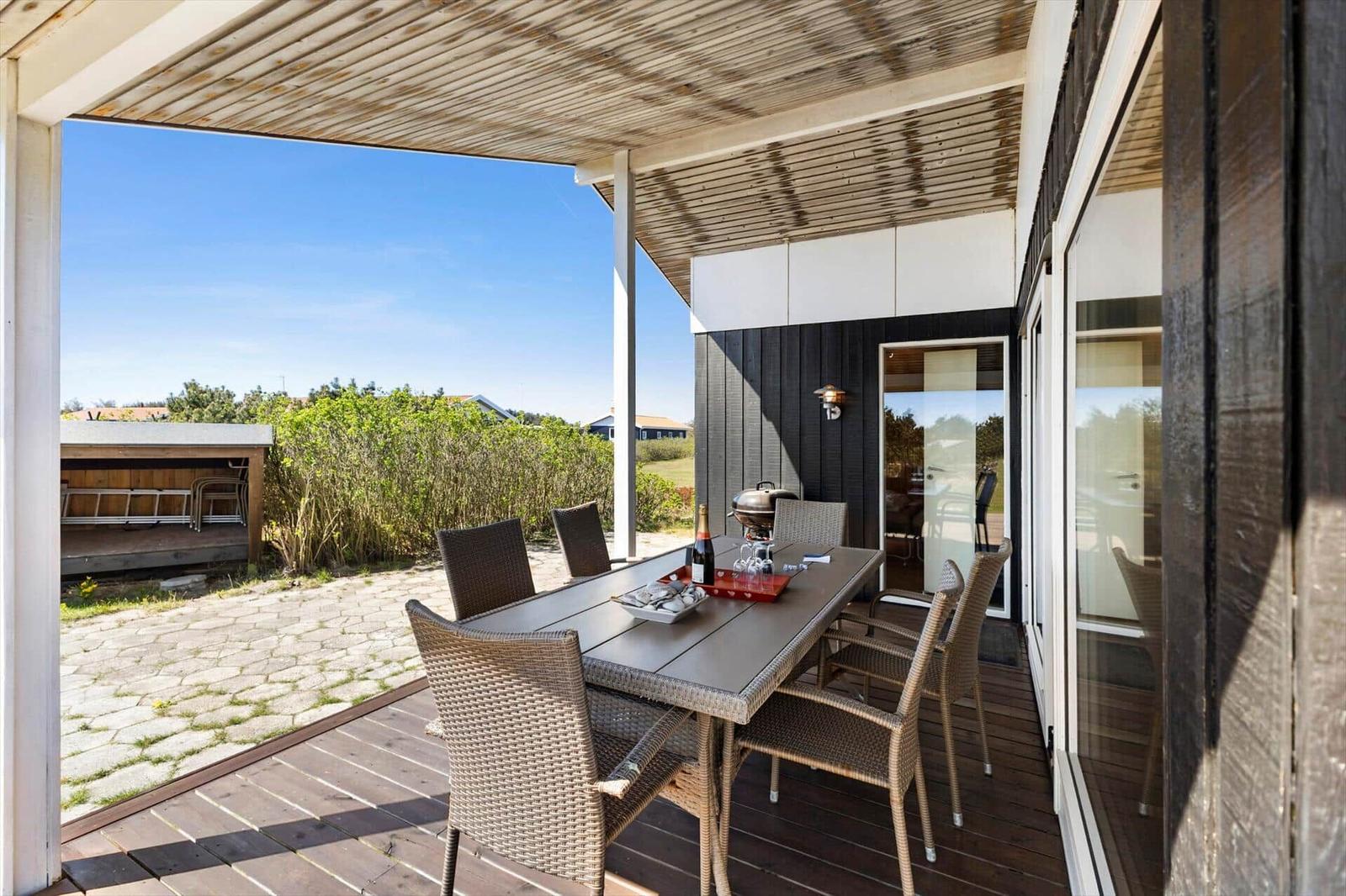 Terrasse mit Esstisch, Stühlen und Grill unter Dach. Blick auf Garten und Himmel.