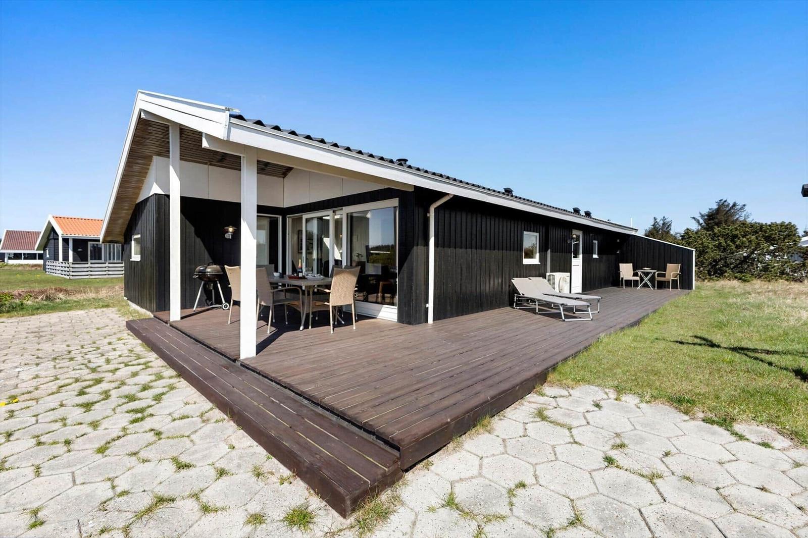 Moderne Villa mit großem Holzterrasse, Grill und Liegen. Blick auf grünen Garten und klaren Himmel.