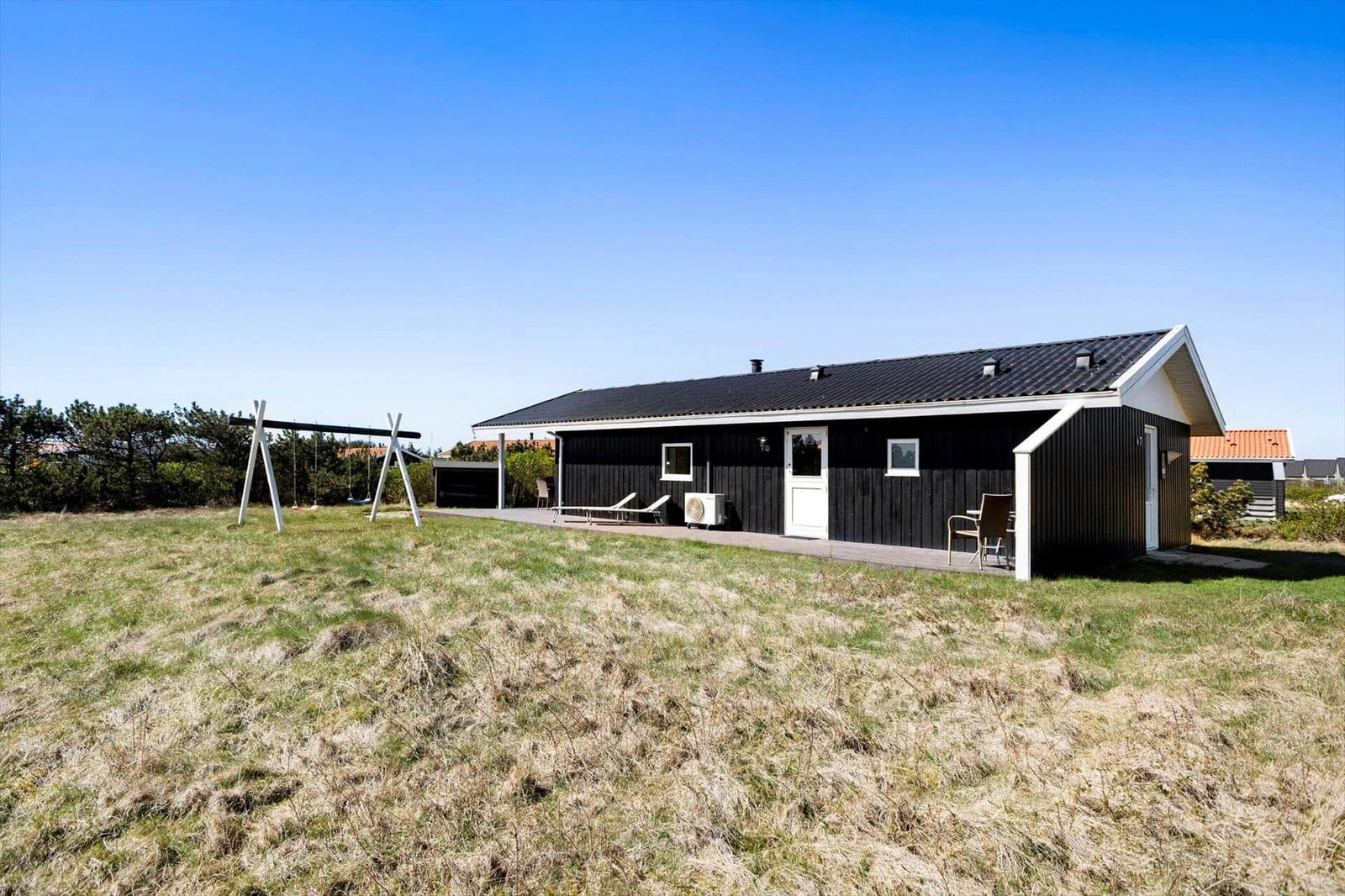Schwarzes Holzhaus mit Terrasse, Liegestühlen und Schaukel im Gras.